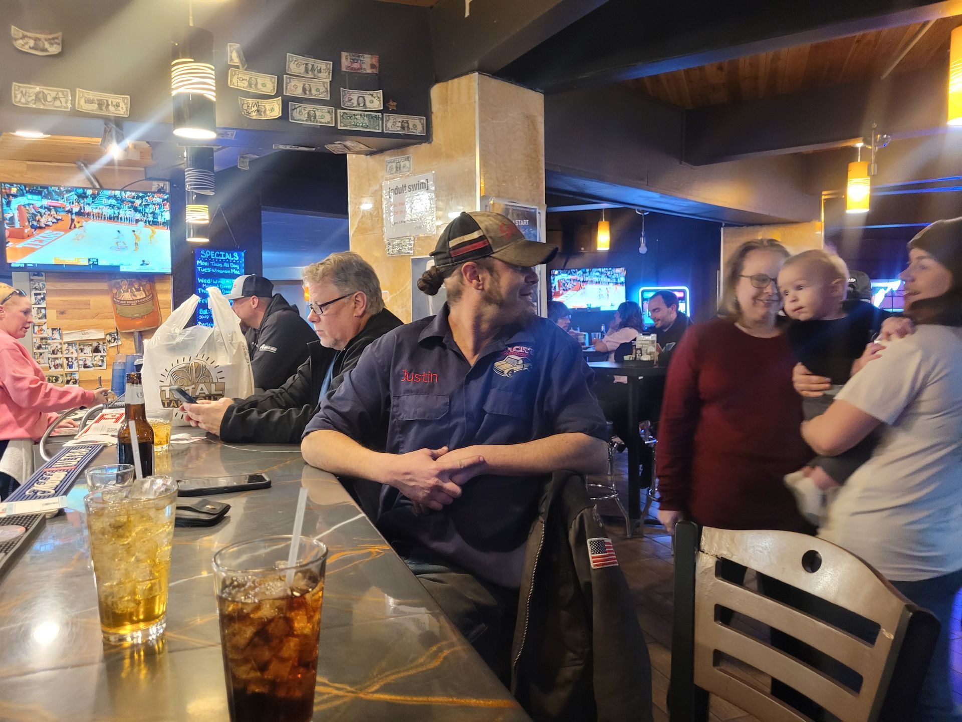 Busy bar with patrons chatting at tables under warm lights and TV screens, with drinks on the counter.