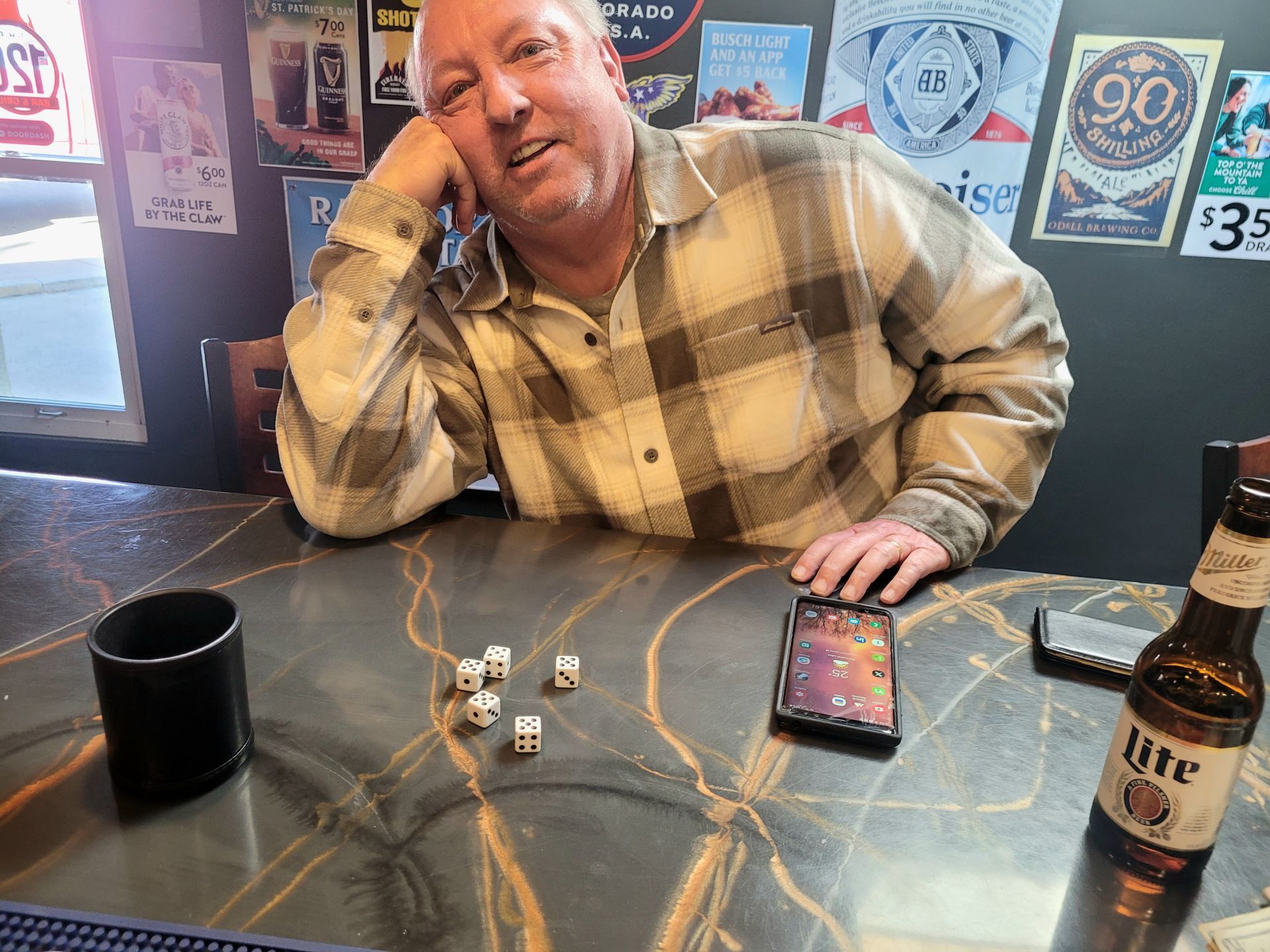 Man smiling at a bar table with dice, a phone, a mug, and a beer bottle.