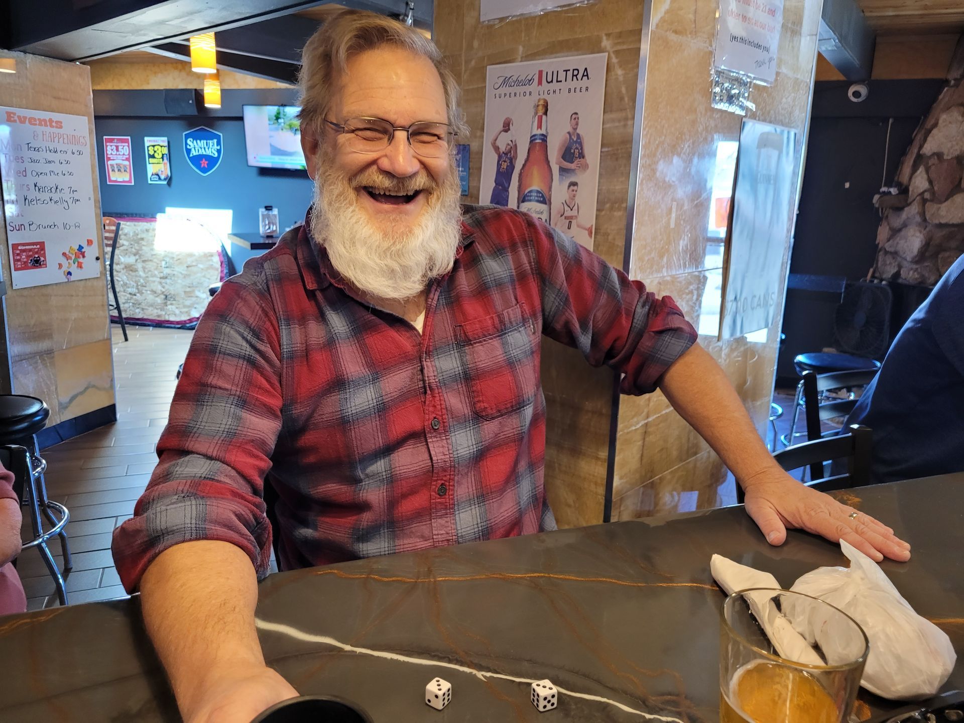 Smiling bearded man in a red plaid shirt leaning on a bar in a rustic indoor setting