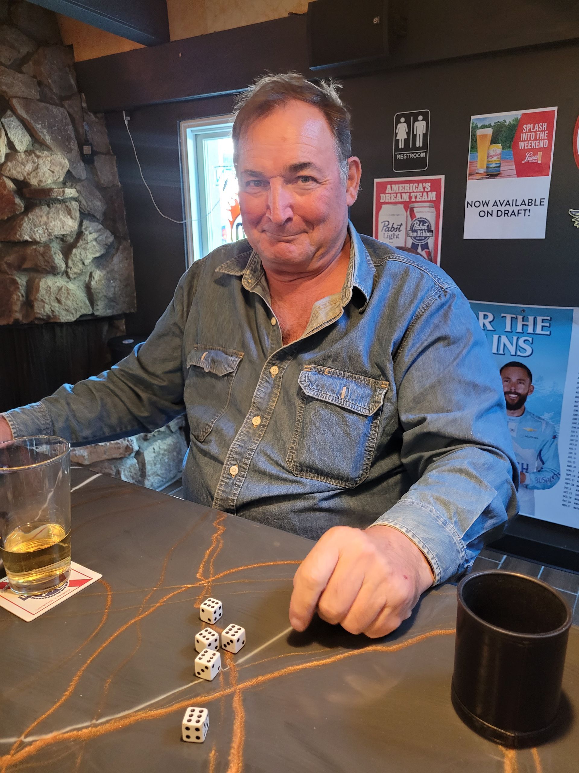 Man seated at a table with dice and drinks in a dim bar, smiling at the camera.