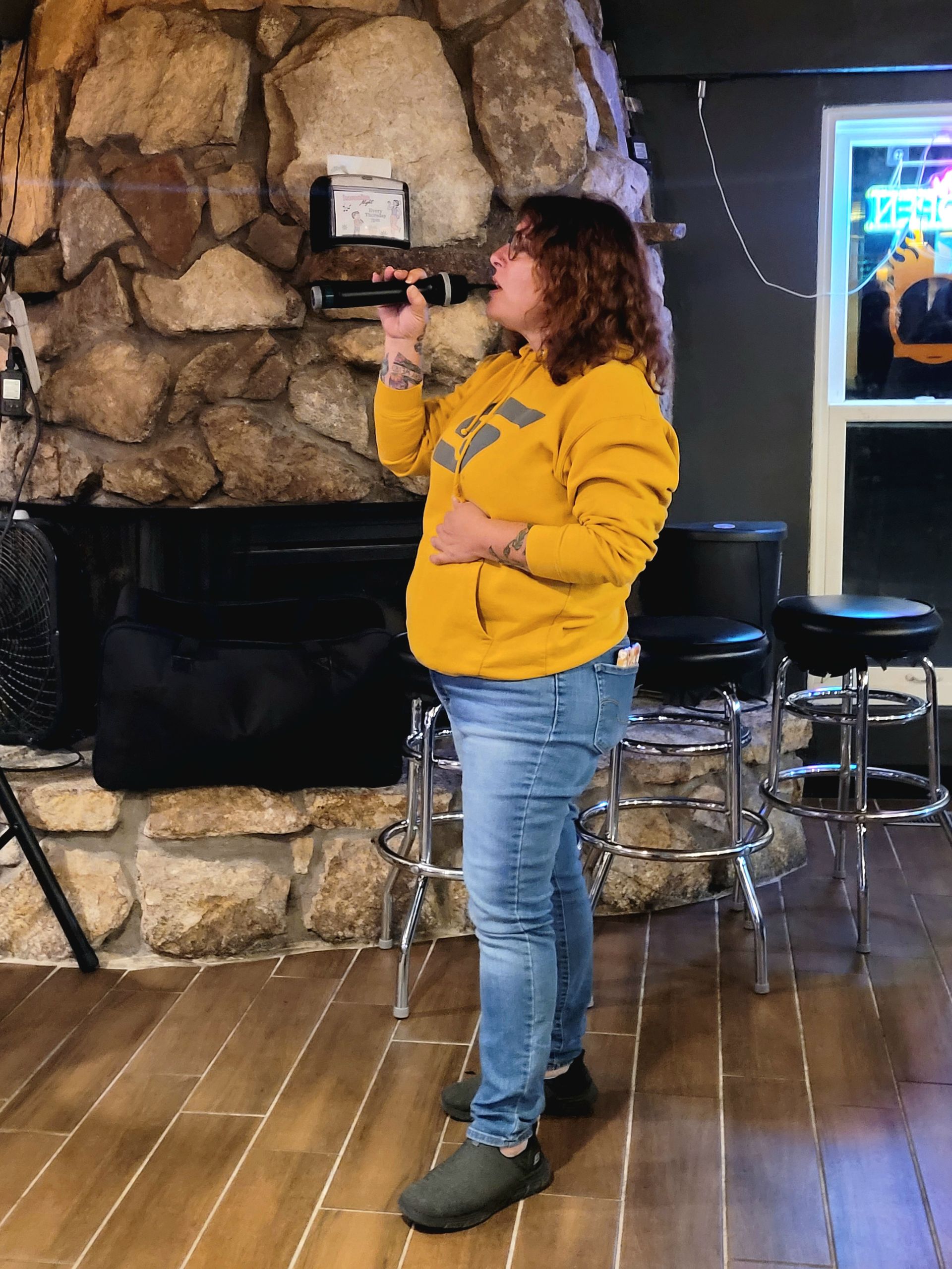 Person in a yellow sweatshirt stands indoors near a stone fireplace and stools, looking to the left.