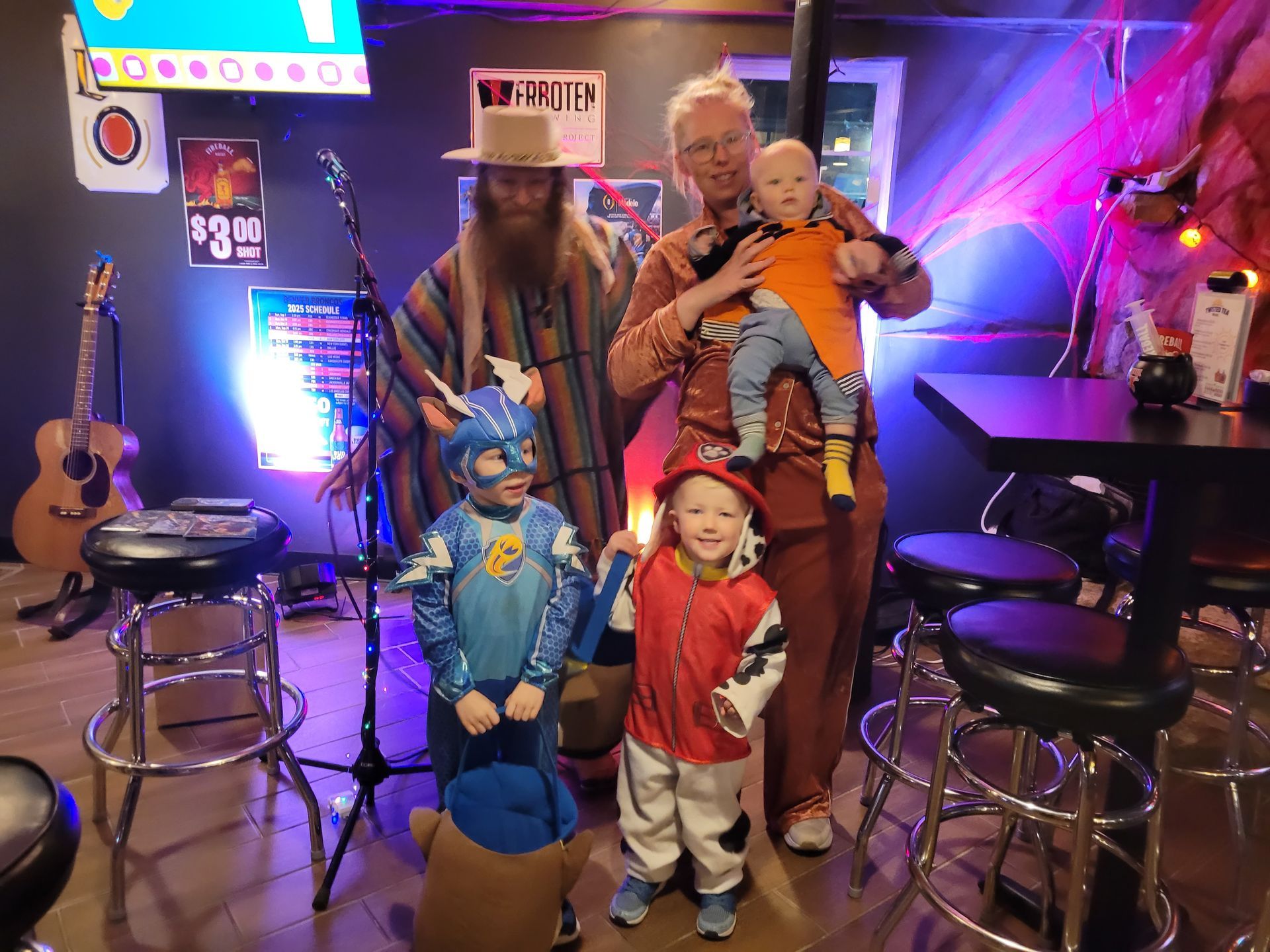 Costumed group posing in a neon-lit bar, with a man holding a baby and two children in superhero outfits