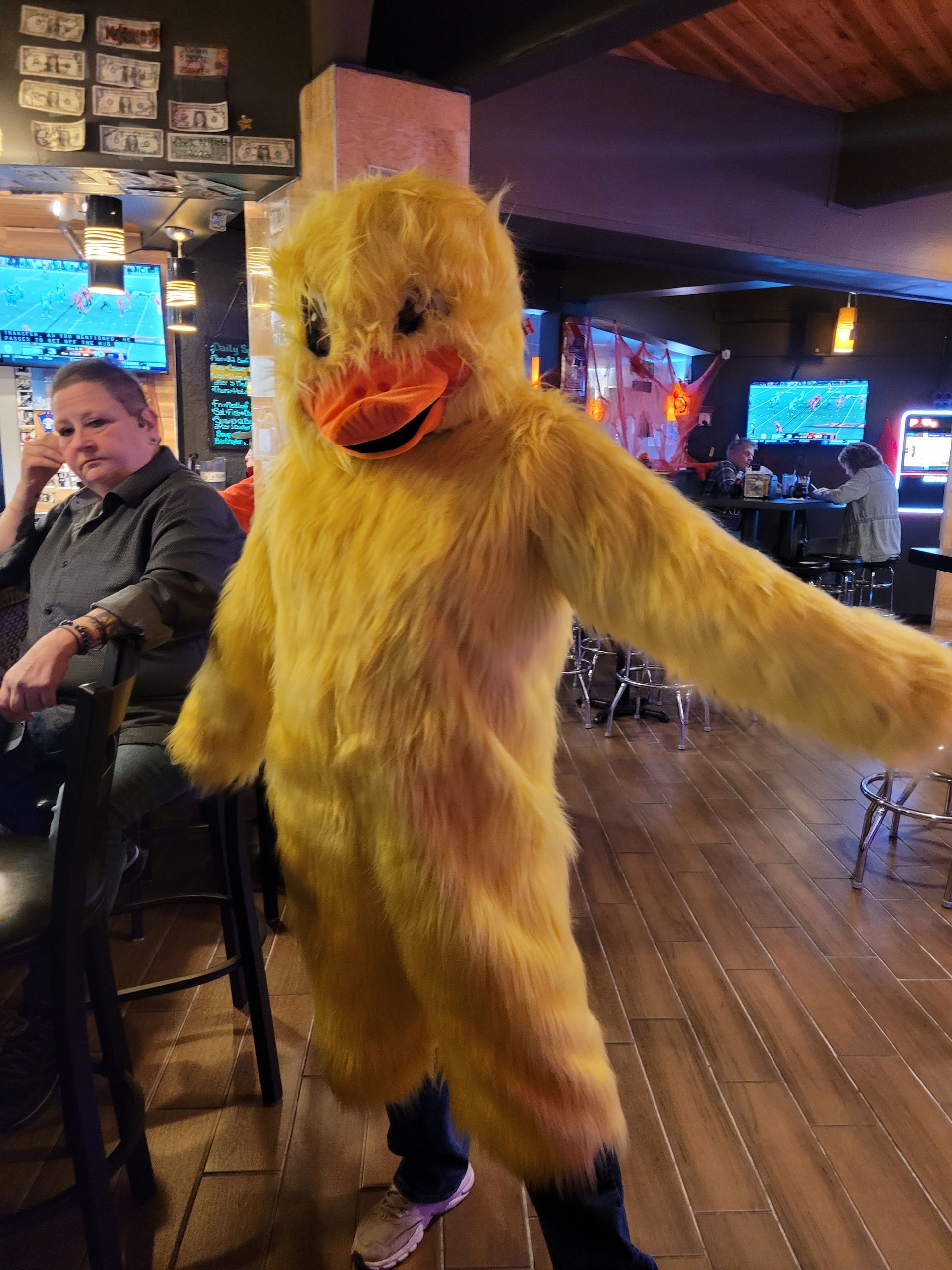 Person in a fluffy yellow duck costume posing in a dimly lit bar or restaurant
