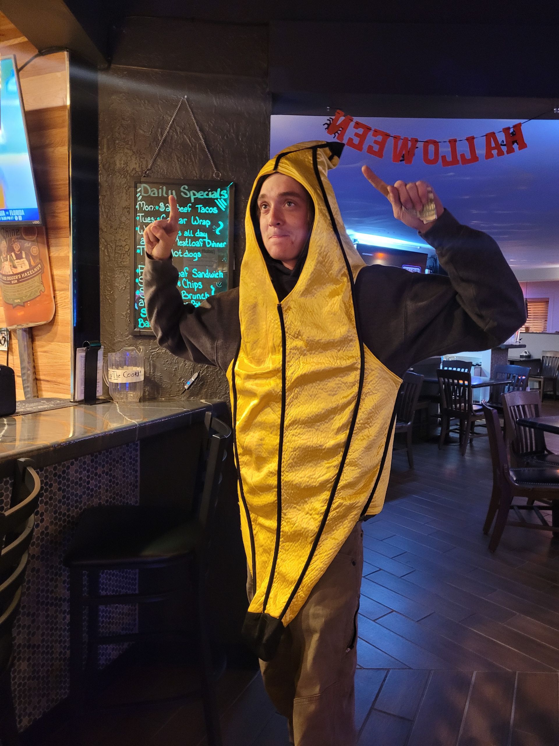 Person in a yellow banana costume posing in a bar or restaurant, arms raised under a neon sign.