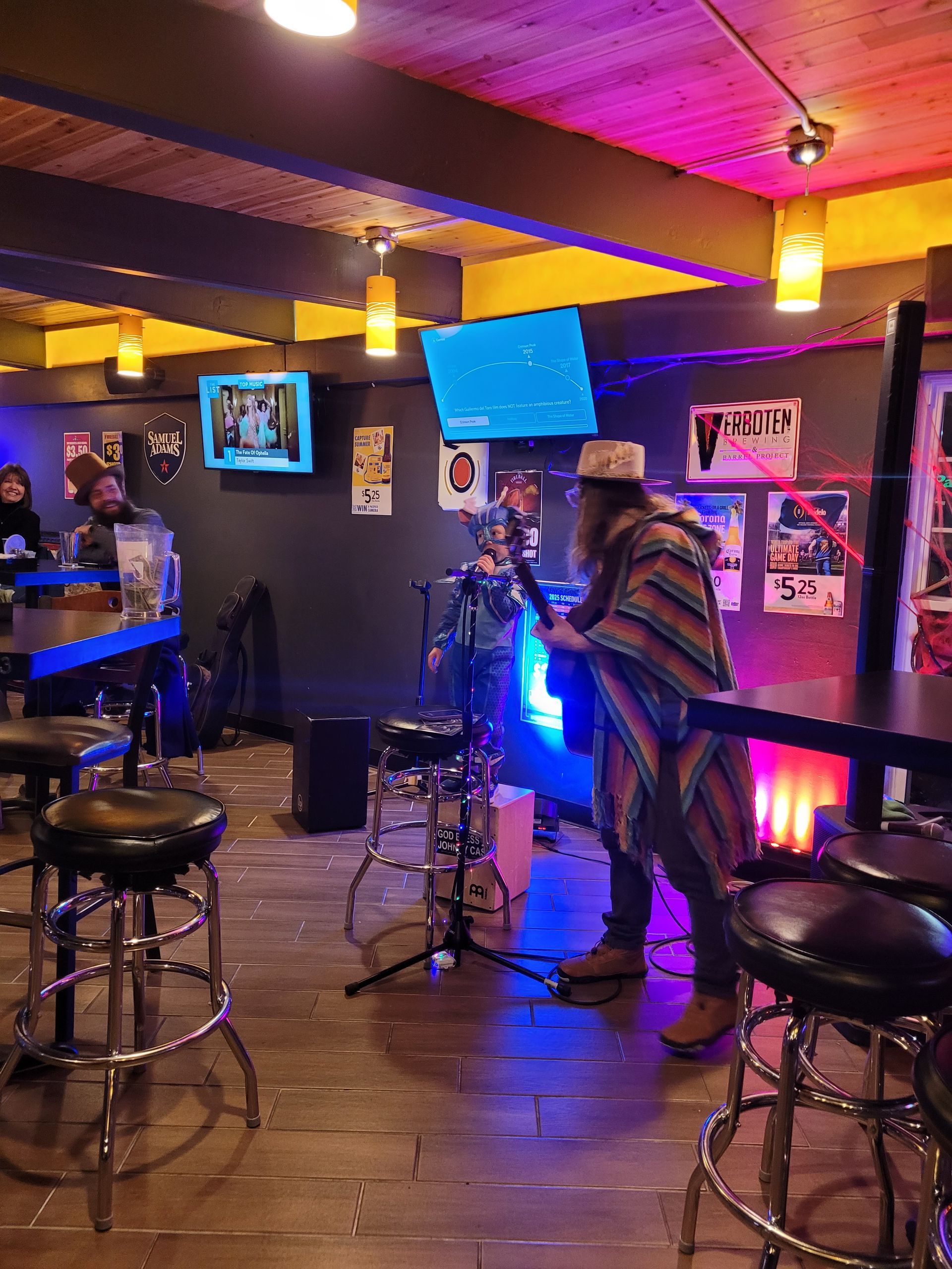 Dimly lit bar interior with stools, neon blue lights, TV screens, and a person playing guitar near the stage