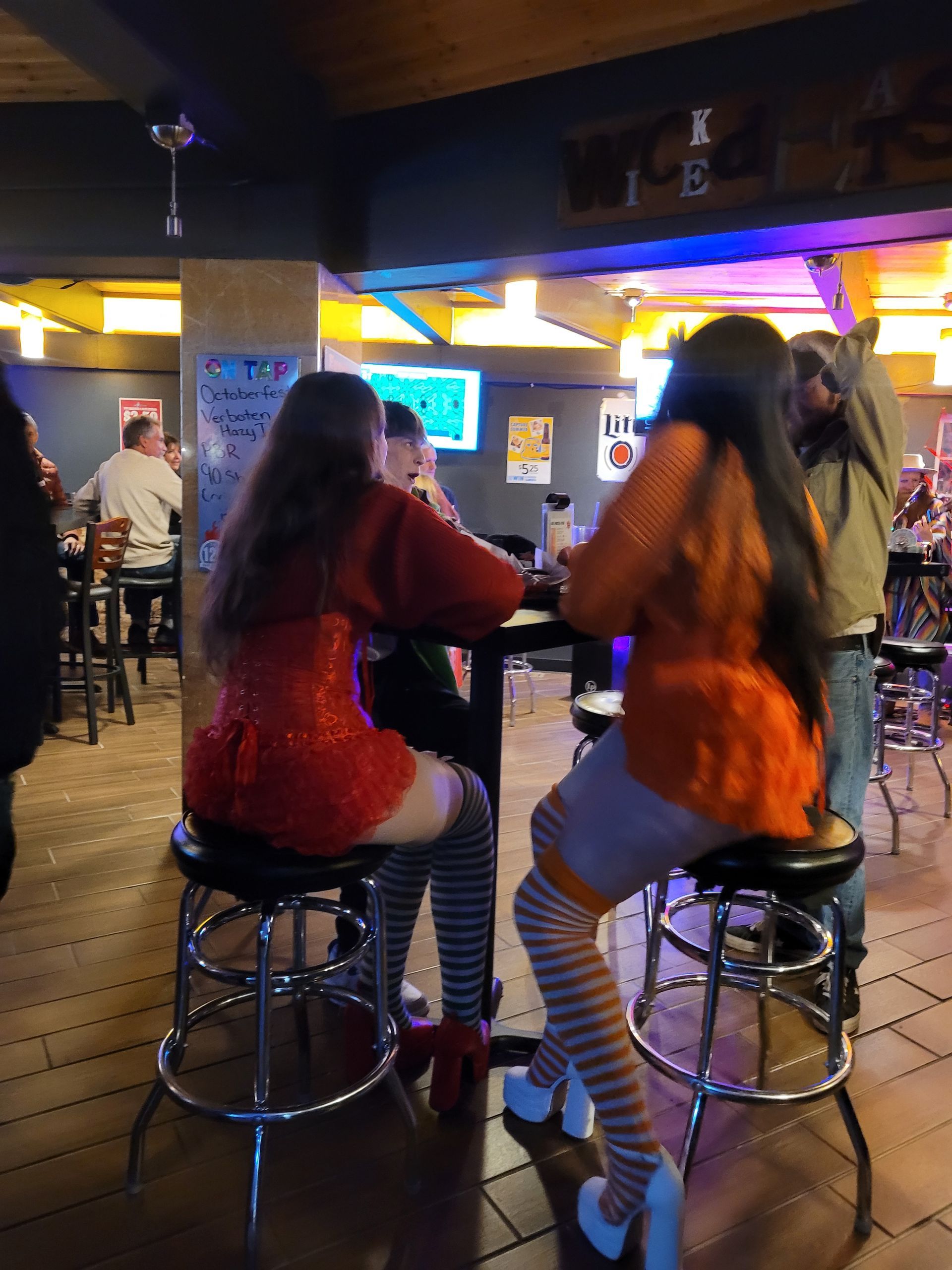 Two people sit on bar stools in a neon-lit arcade bar, facing game screens and chatting.