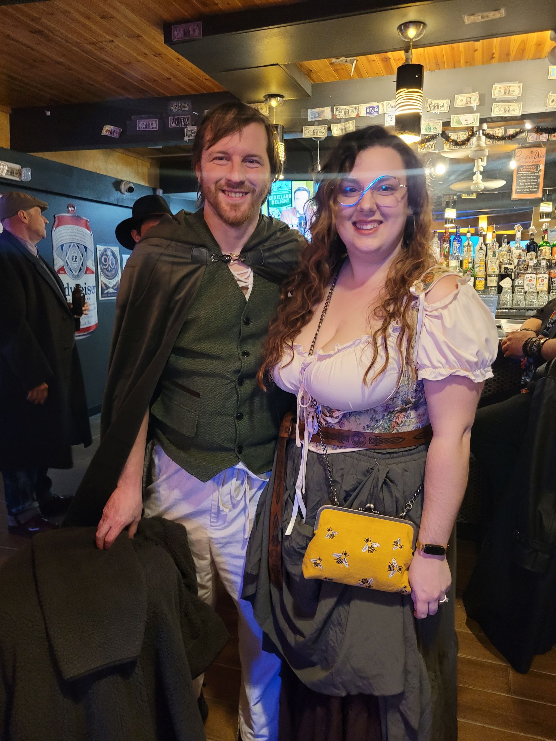 Two people smiling in a dim bar, standing close together under warm lights; one wears a green shirt, the other a purple dress.