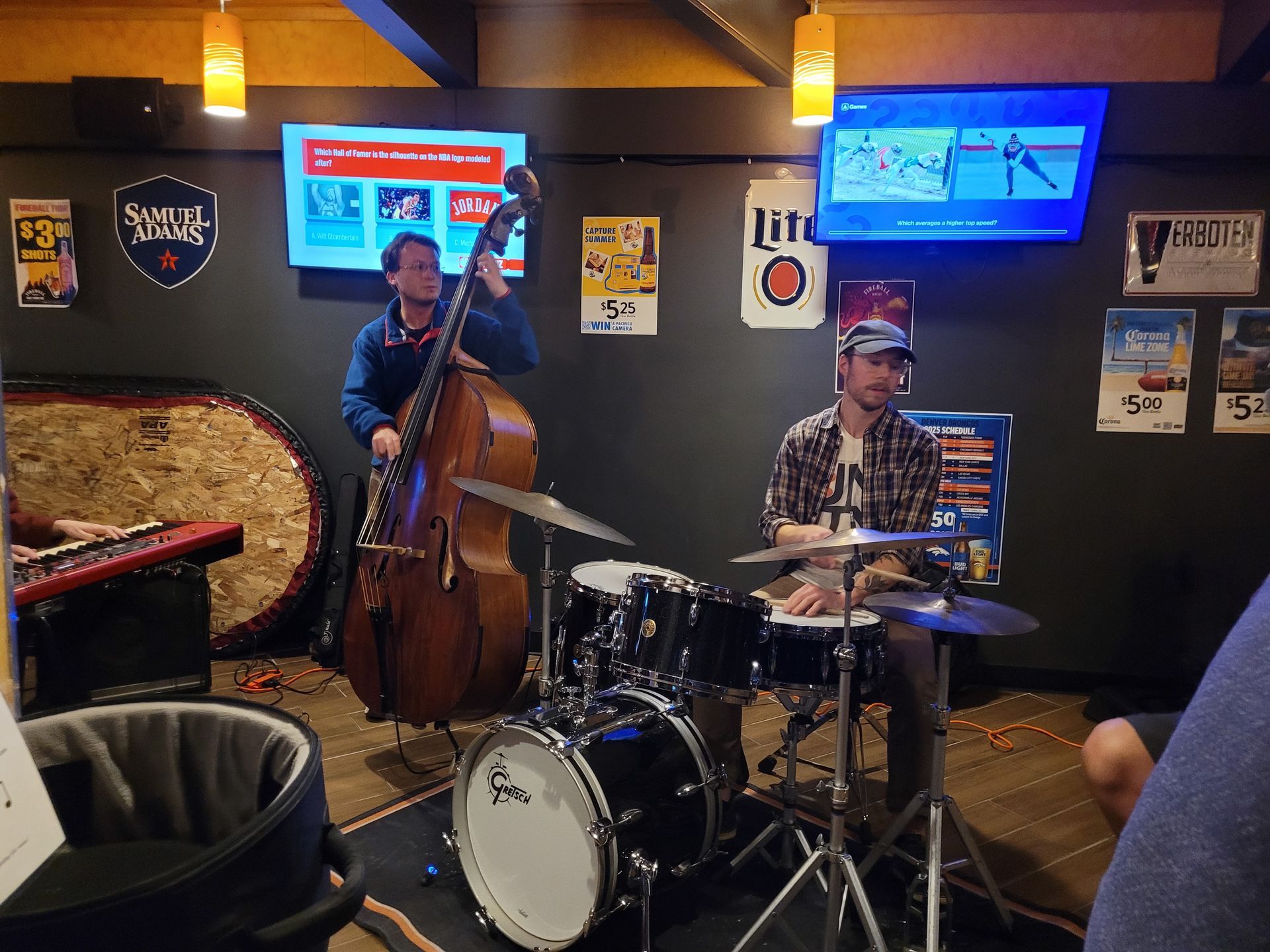 Live jazz duo playing bass and drums in a dim bar with TV screens on the wall.