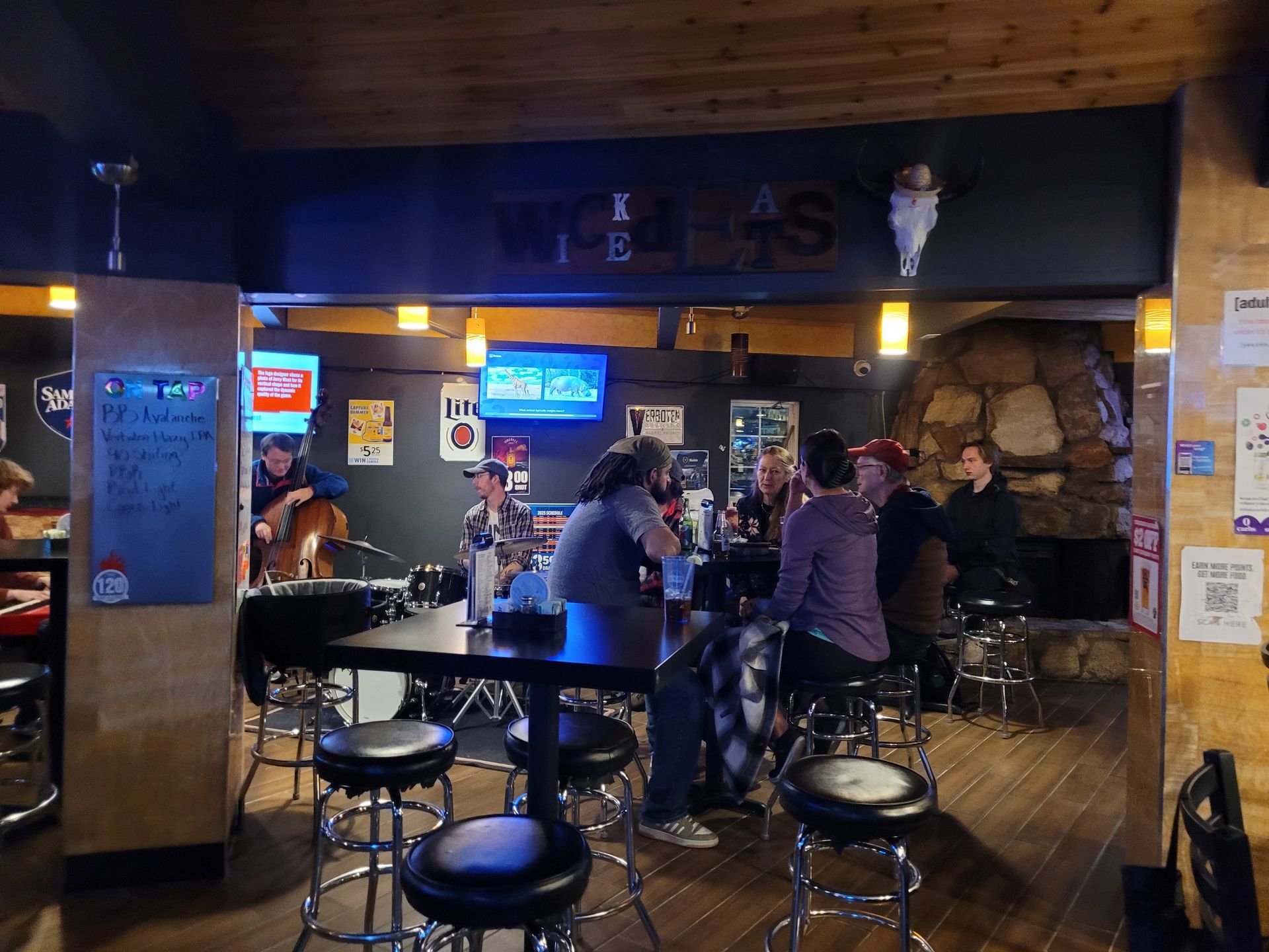 Dimly lit bar interior with patrons seated at high tables and stools near TVs and stone wall.