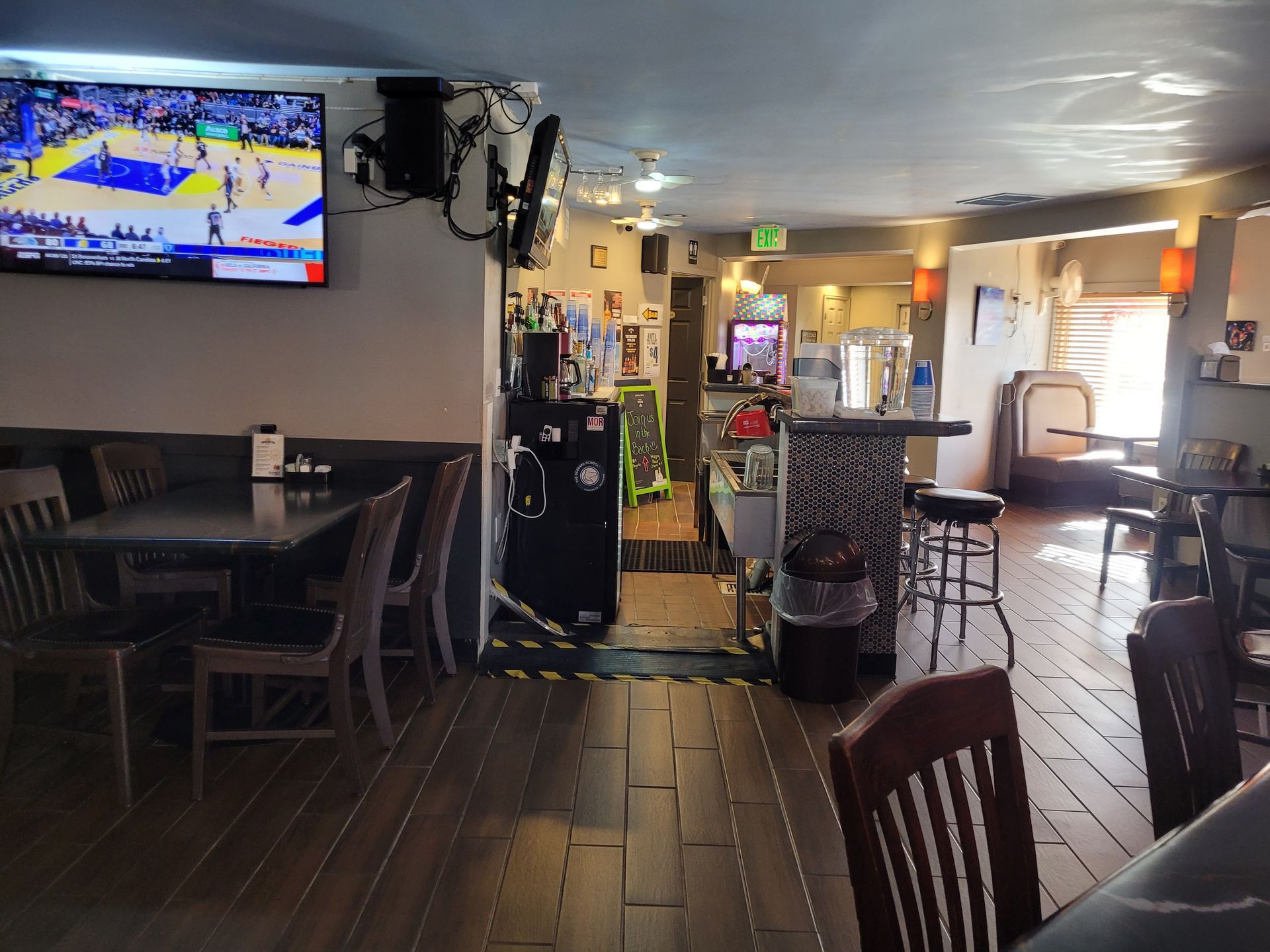 Dim restaurant dining room with TV showing a basketball game, wooden tables, and bar stools near the counter.