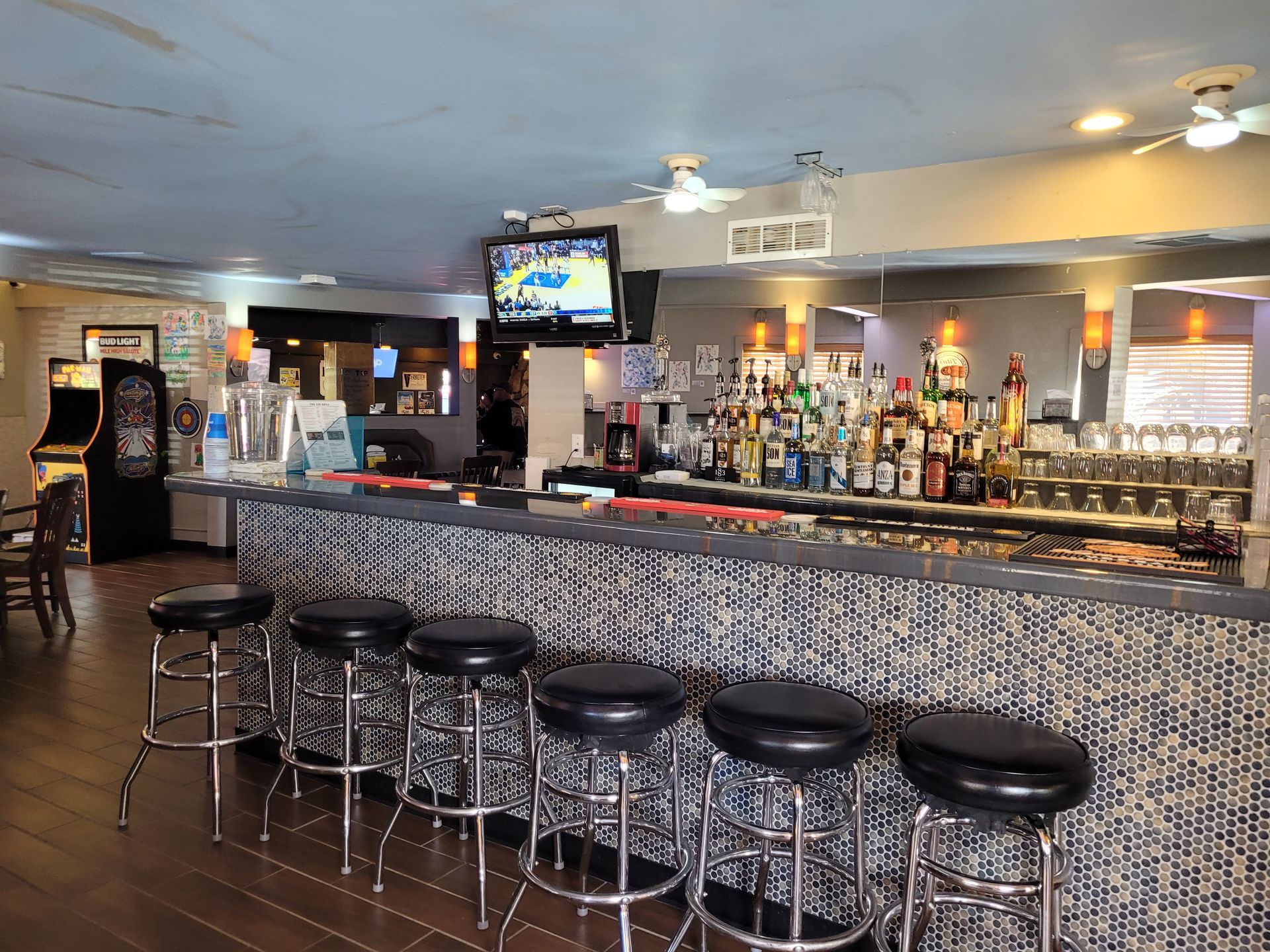 Empty bar interior with stools, patterned counter, liquor bottles, and a TV overhead.