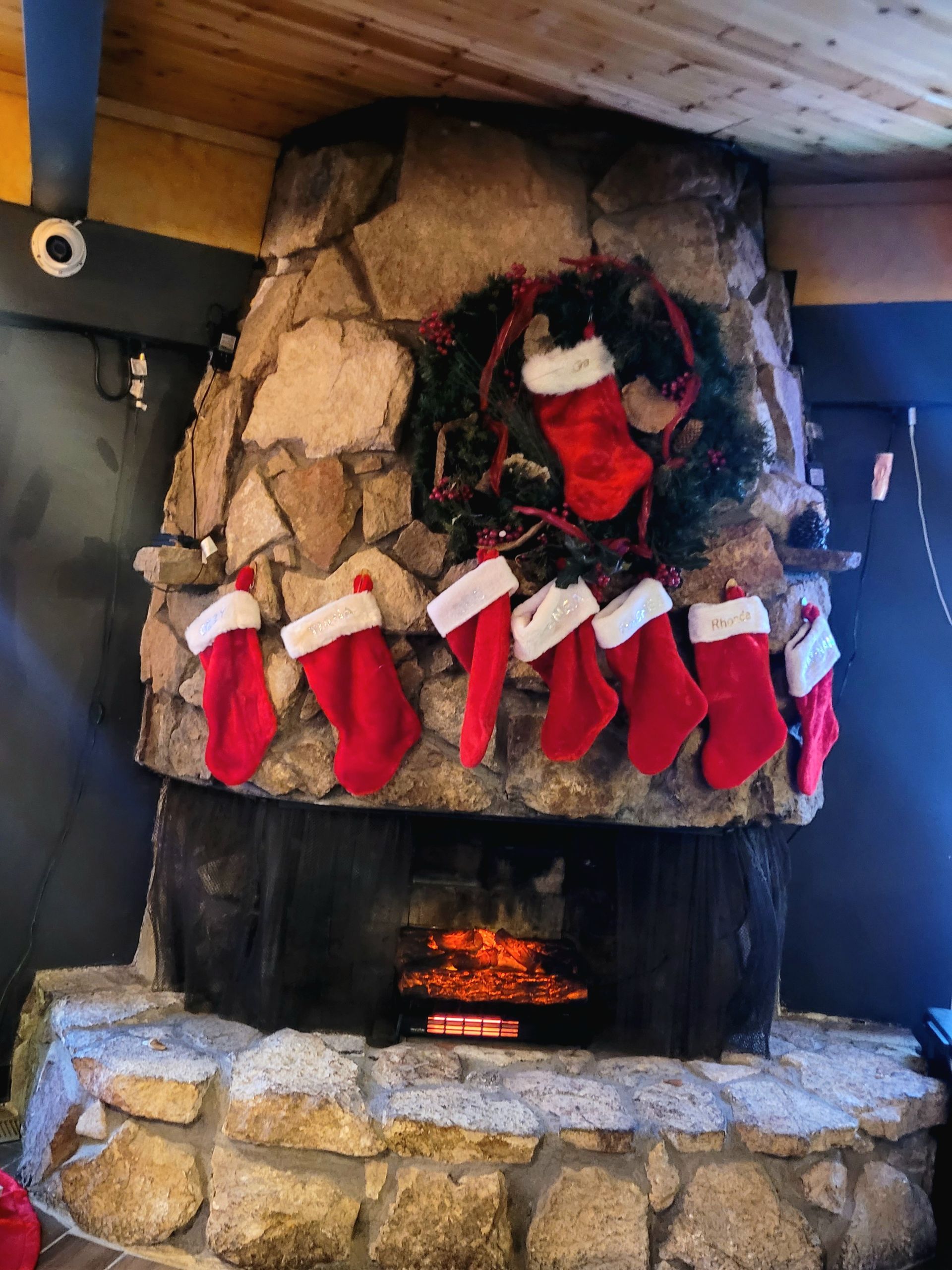 Christmas stockings and a wreath hung on a rustic stone fireplace with a small fire burning below