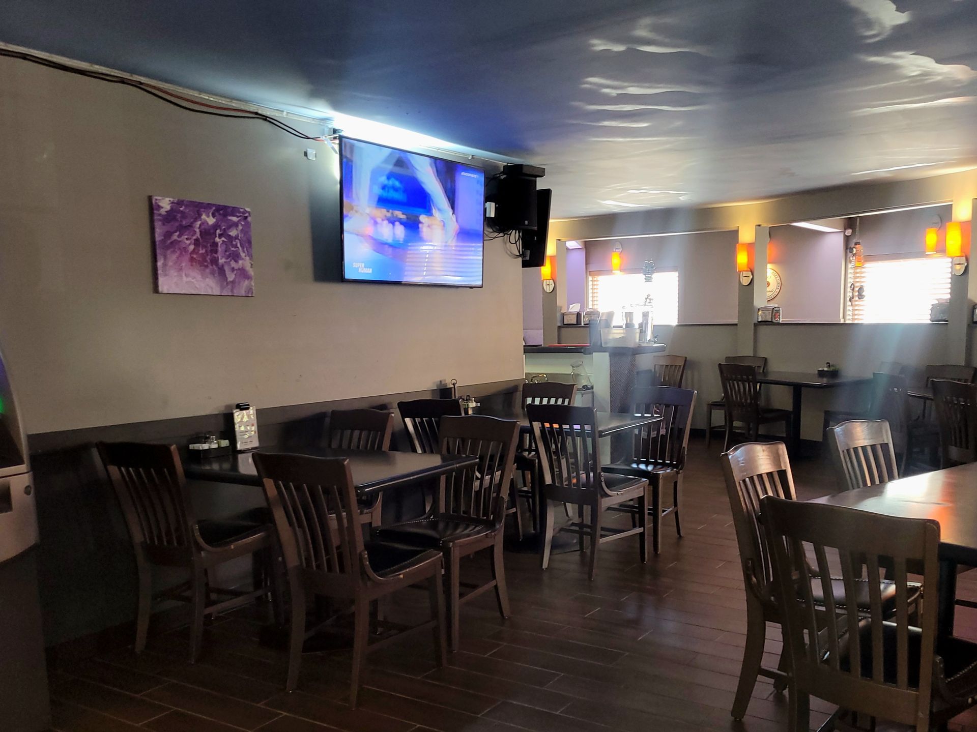 Empty restaurant dining room with dark tables and chairs, wall TV, and bright windows in the back.