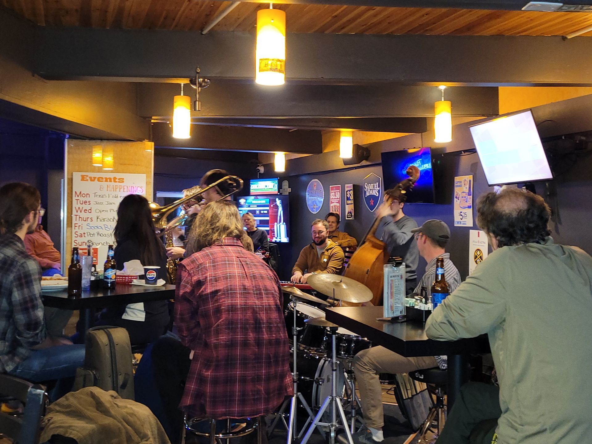 People seated at a bar in a dimly lit venue, with hanging lights and TVs in the background.