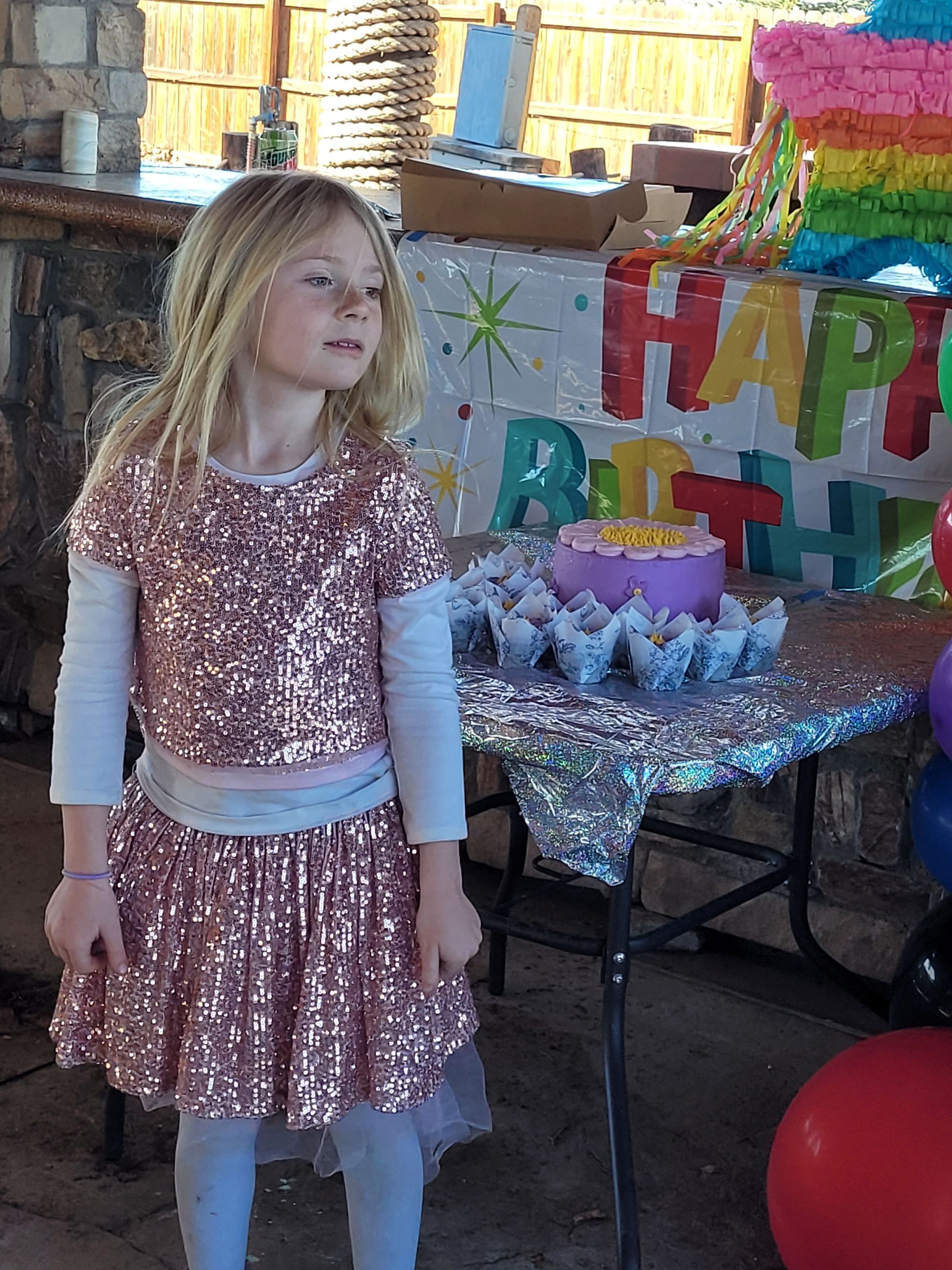 Girl in a sparkly dress standing beside a birthday party table with cupcakes and colorful decorations.