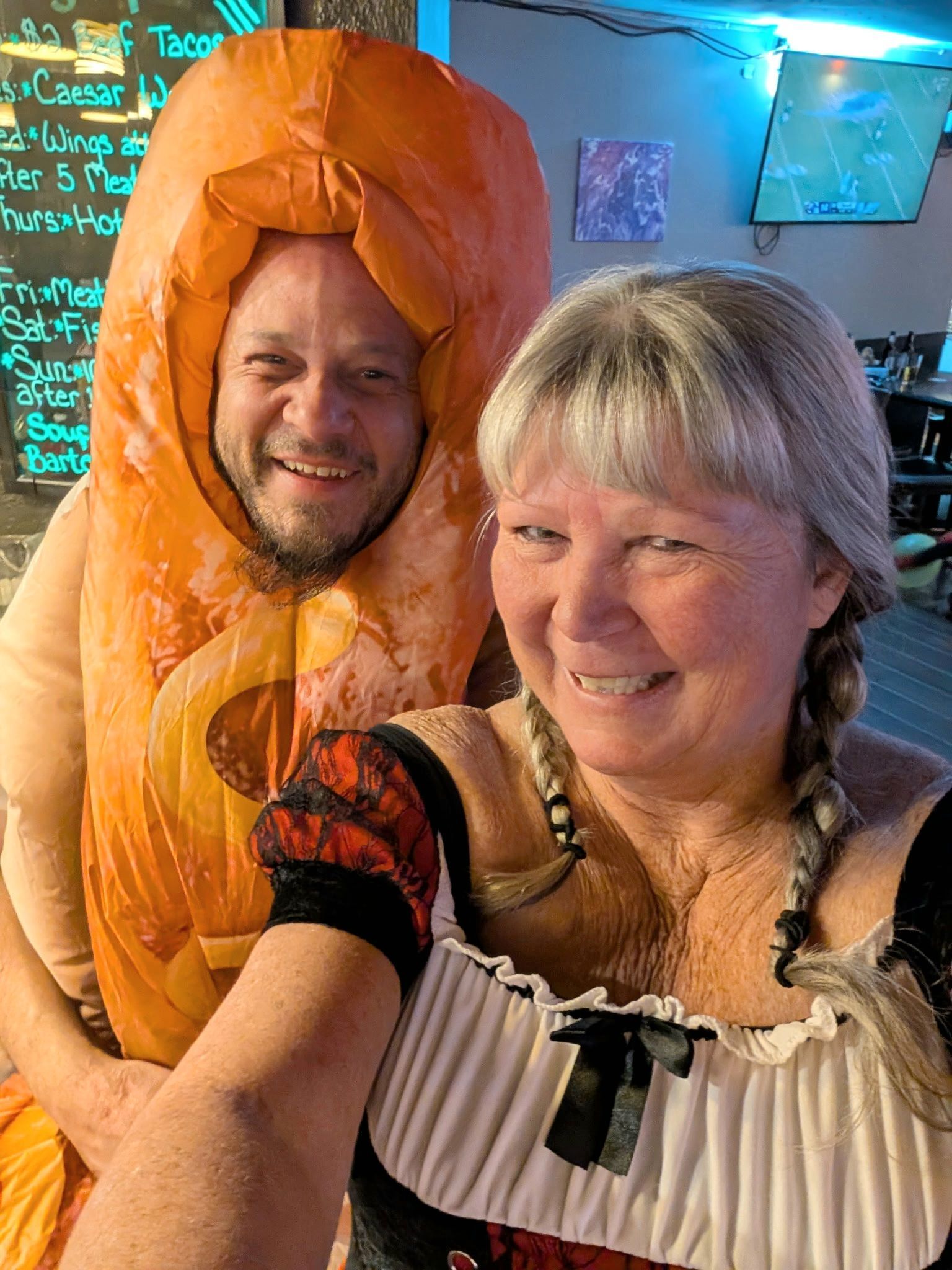 Two smiling people posing indoors, one wearing an orange costume hood and the other in a black-and-white top.