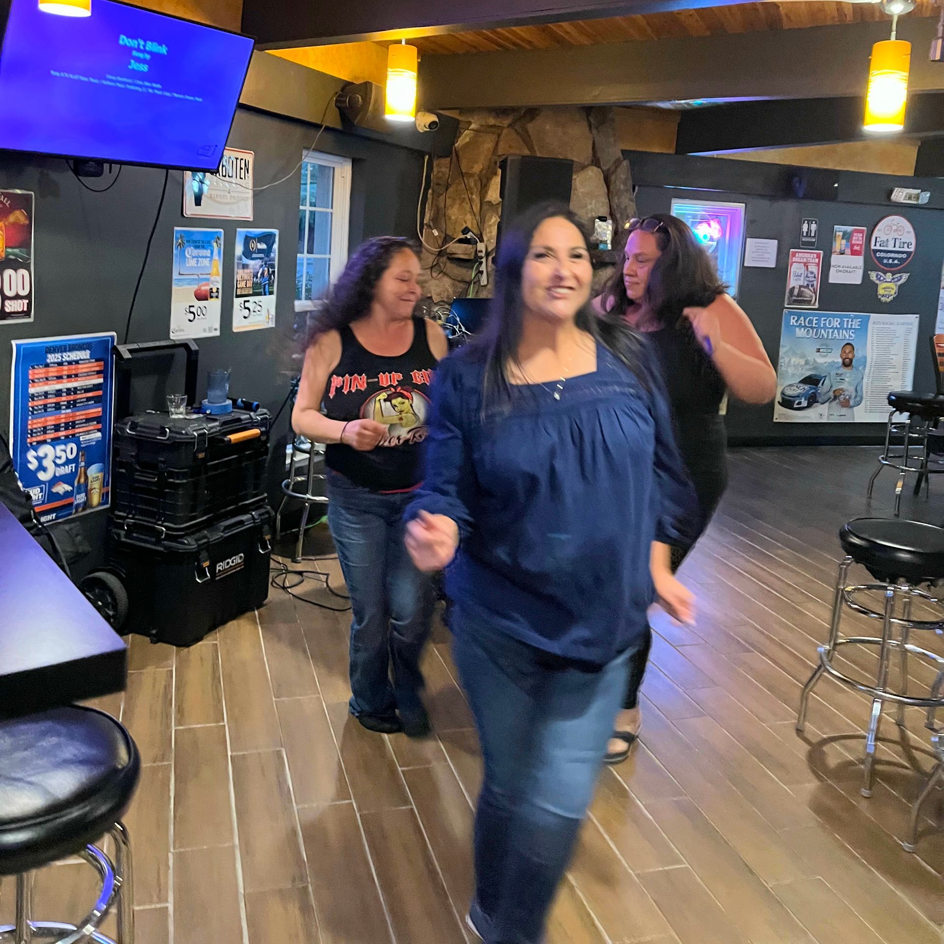 Two women walking toward the camera in a dimly lit bar with wood floors and neon signs