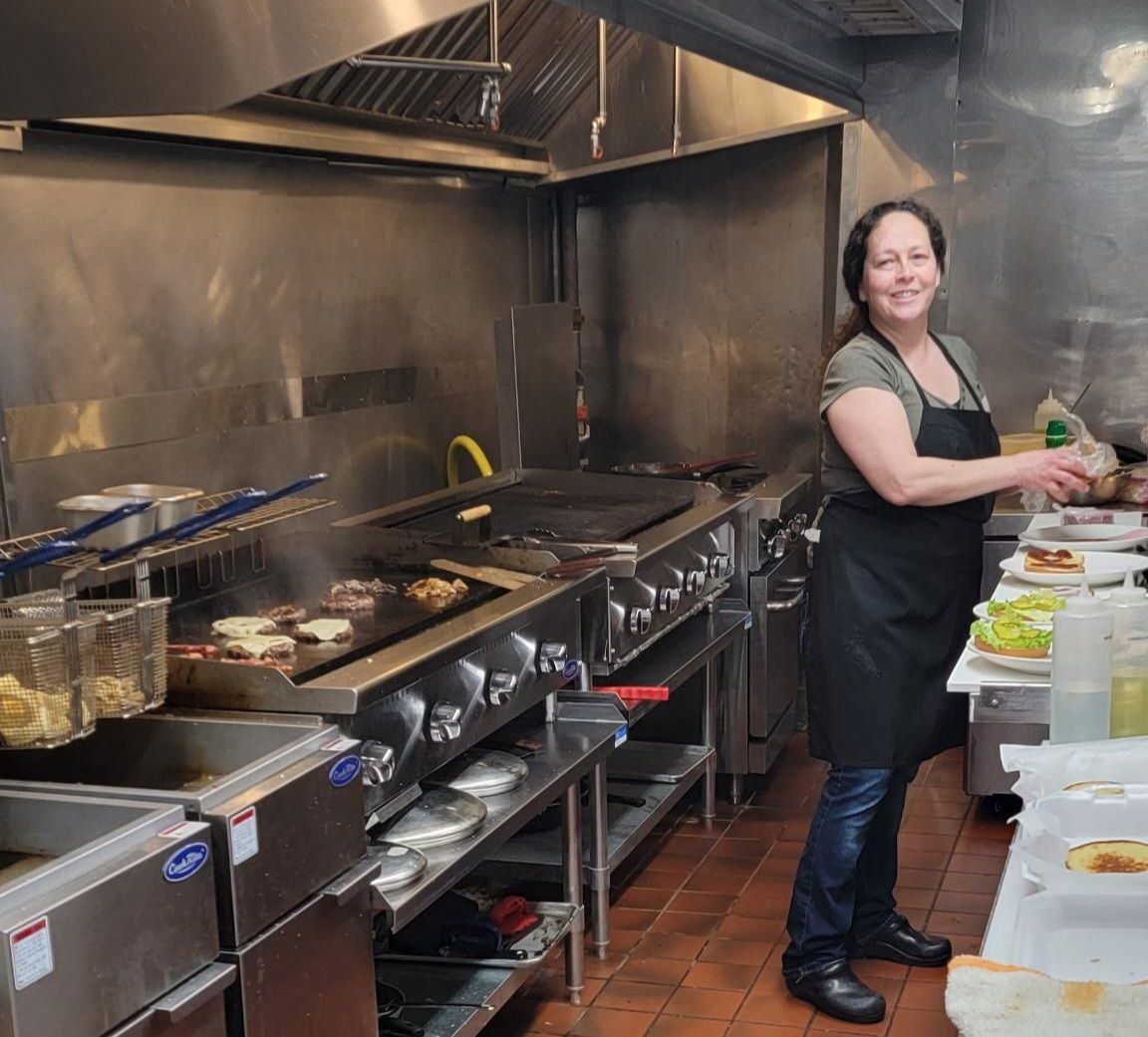 Chef smiling in a stainless-steel kitchen, plating food beside a grill and prep table