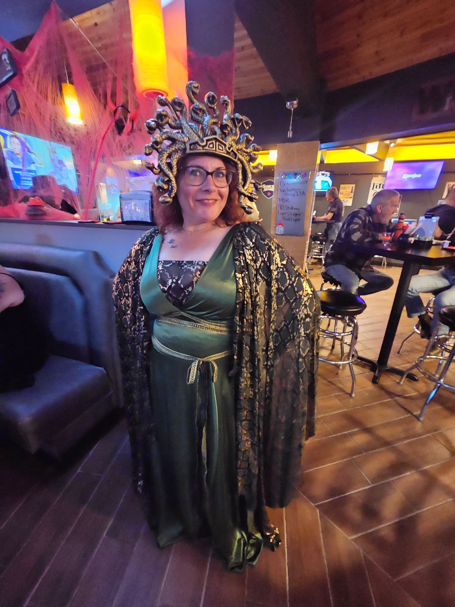 Person in a green gown and jeweled headpiece standing in a dimly lit bar with colorful lights.