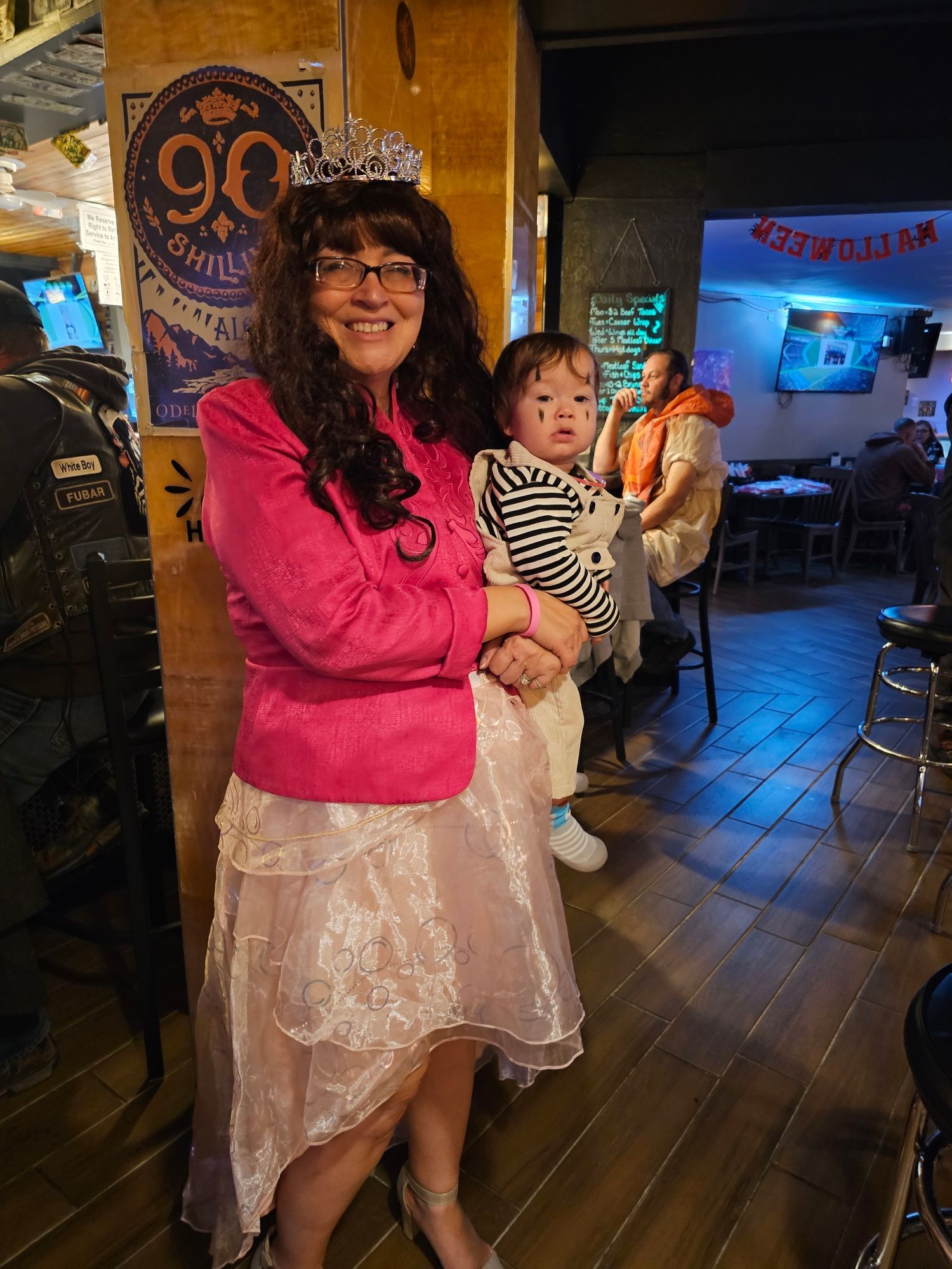 Woman in bright pink jacket and white lace skirt standing indoors, smiling beside a child in a striped shirt.