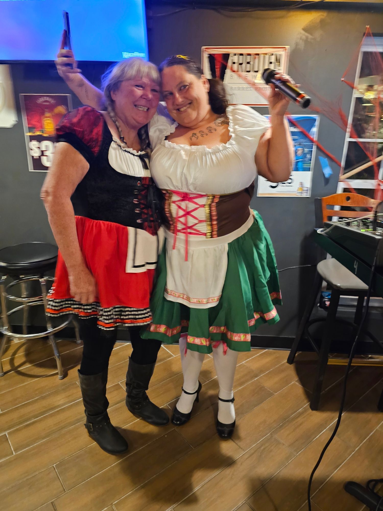 Two women in dirndls pose smiling with drinks at a lively indoor event, one holding up a beer bottle.