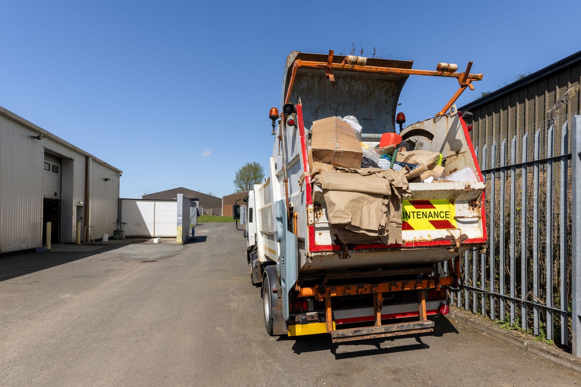 Garbage truck loaded with trash parked in an industrial alley beside buildings and a fence