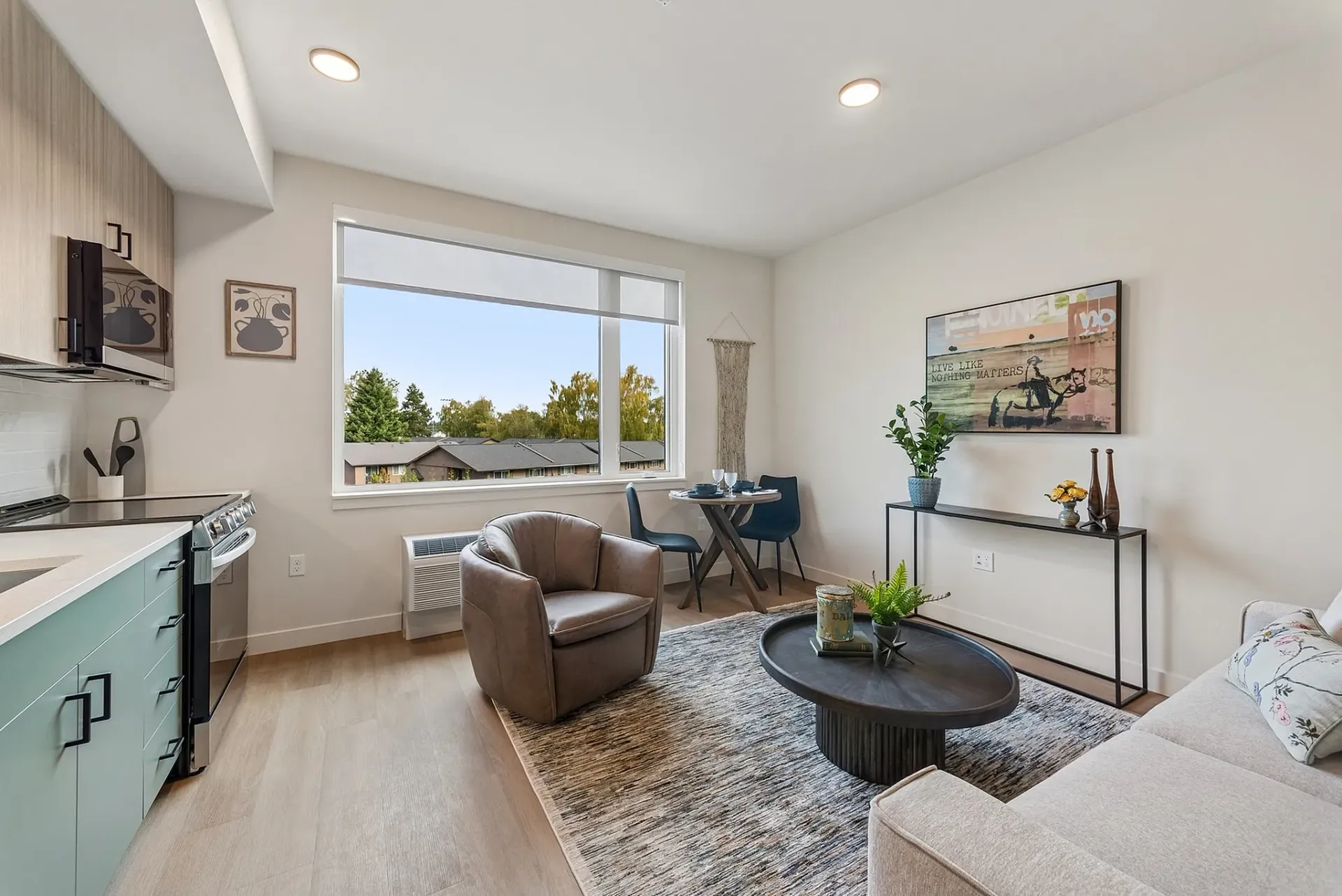Living room with a kitchen area, modern furniture, and a large window with a view of trees and houses.