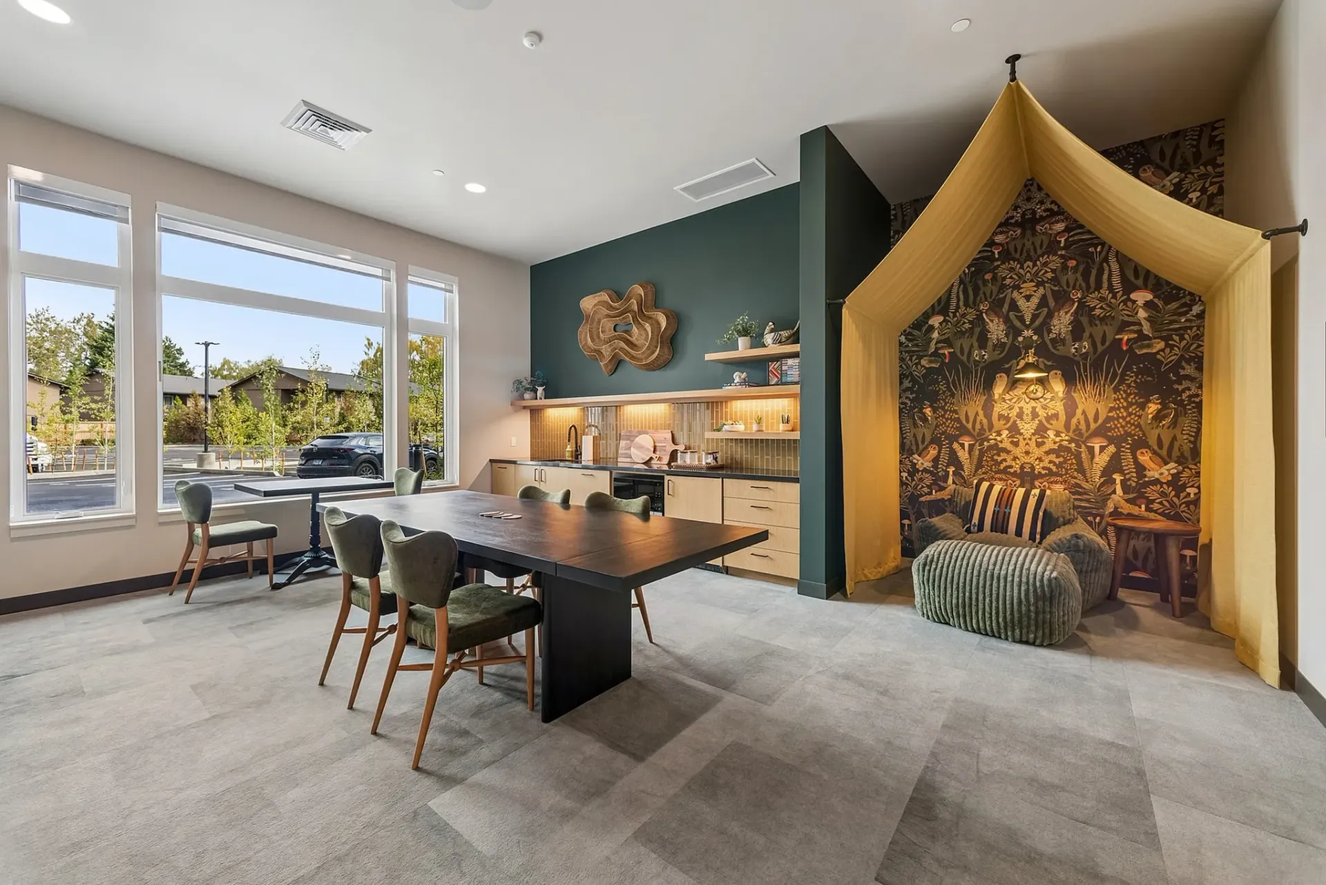 Community lounge area with a large table, kitchen, and reading nook with unique wallpaper.