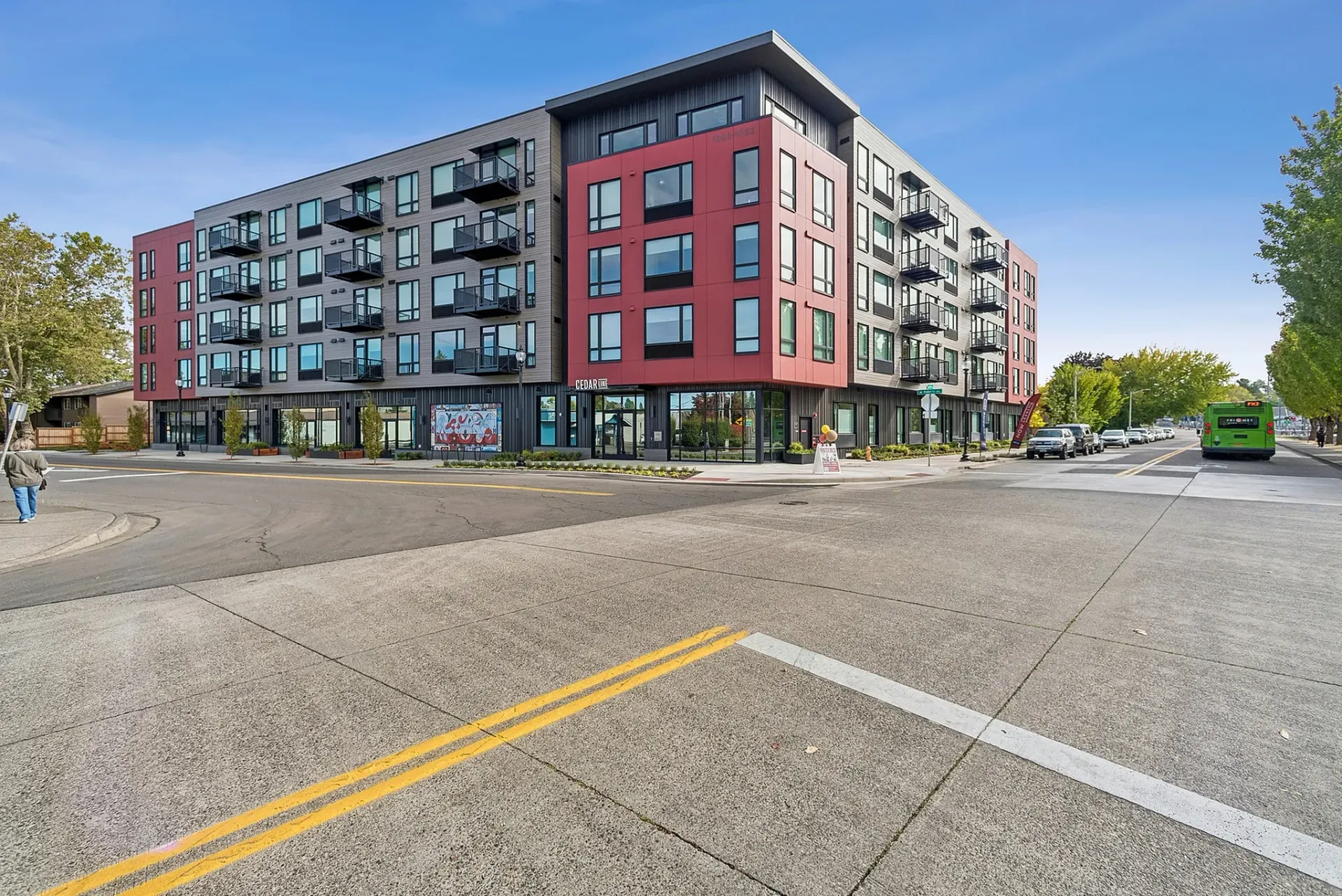 Modern apartment building exterior with red and gray accents, balconies, and ground-floor retail spaces.