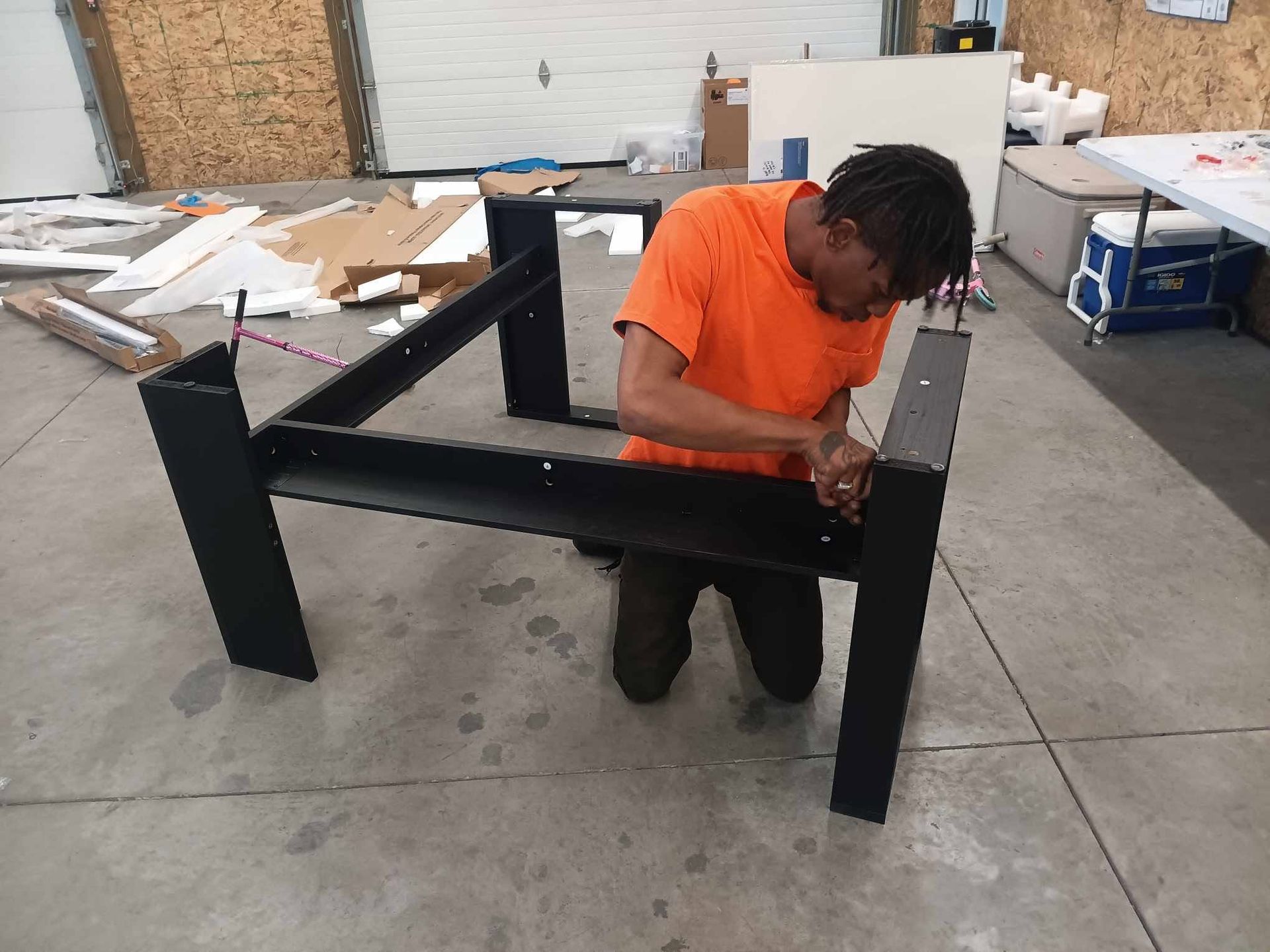 Person in an orange shirt assembling a black metal frame in a workshop.