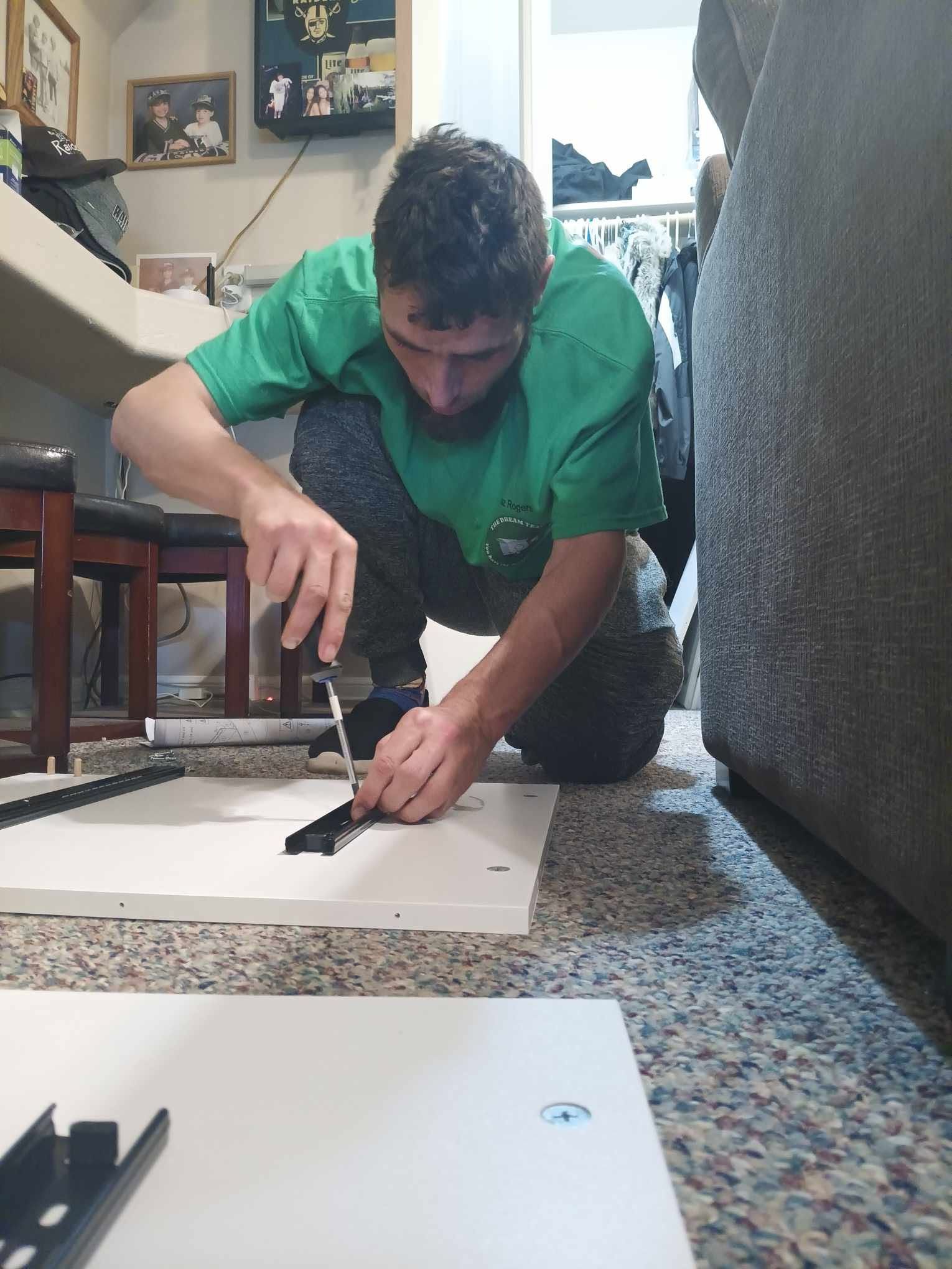 Person in a green shirt measuring paper on the floor beside a couch, using a ruler and knife.