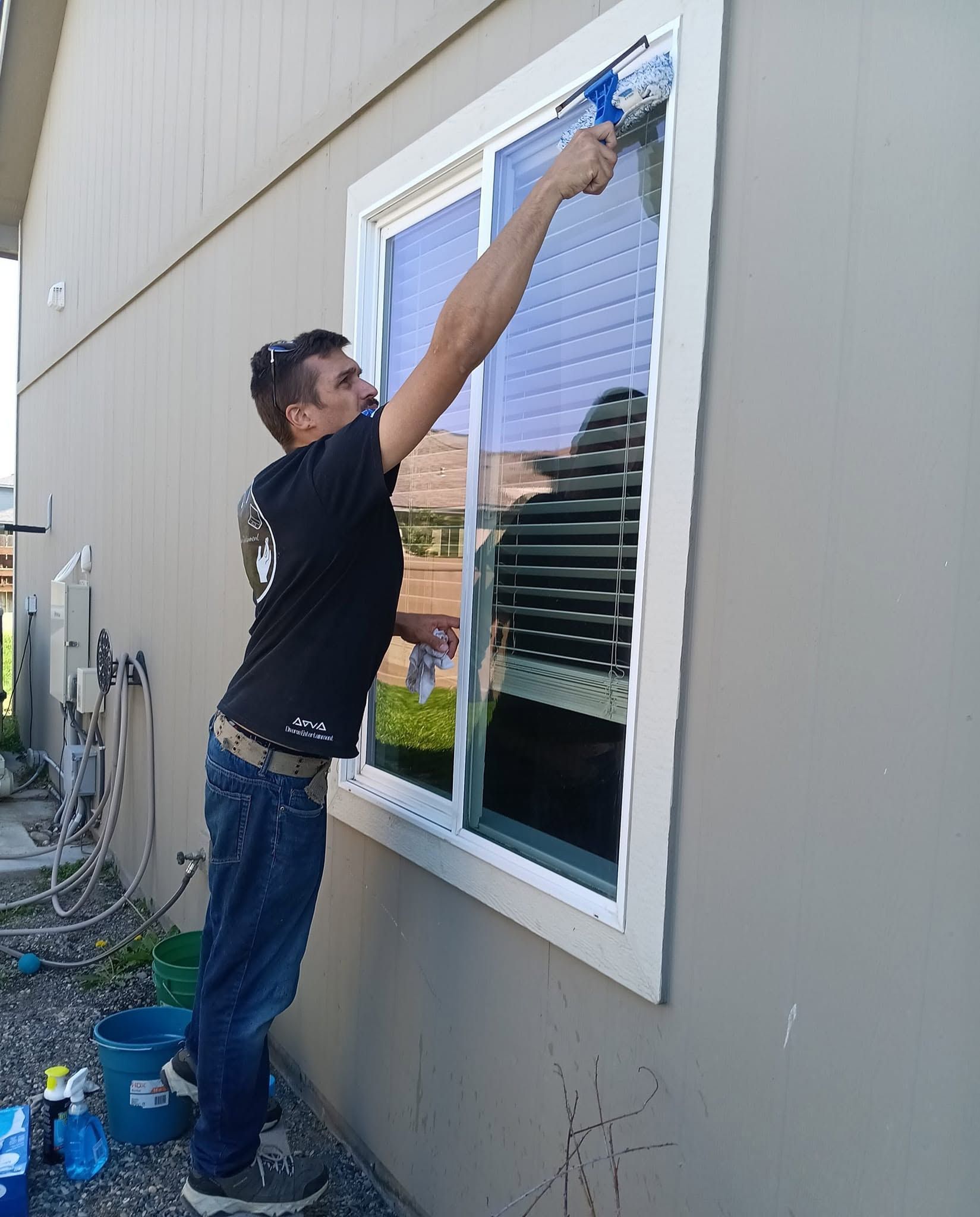Man cleaning an exterior window on a house with a blue cloth, standing on a bucket step.