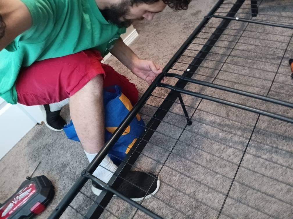 Person assembling a black metal bed frame on carpet with a power drill nearby