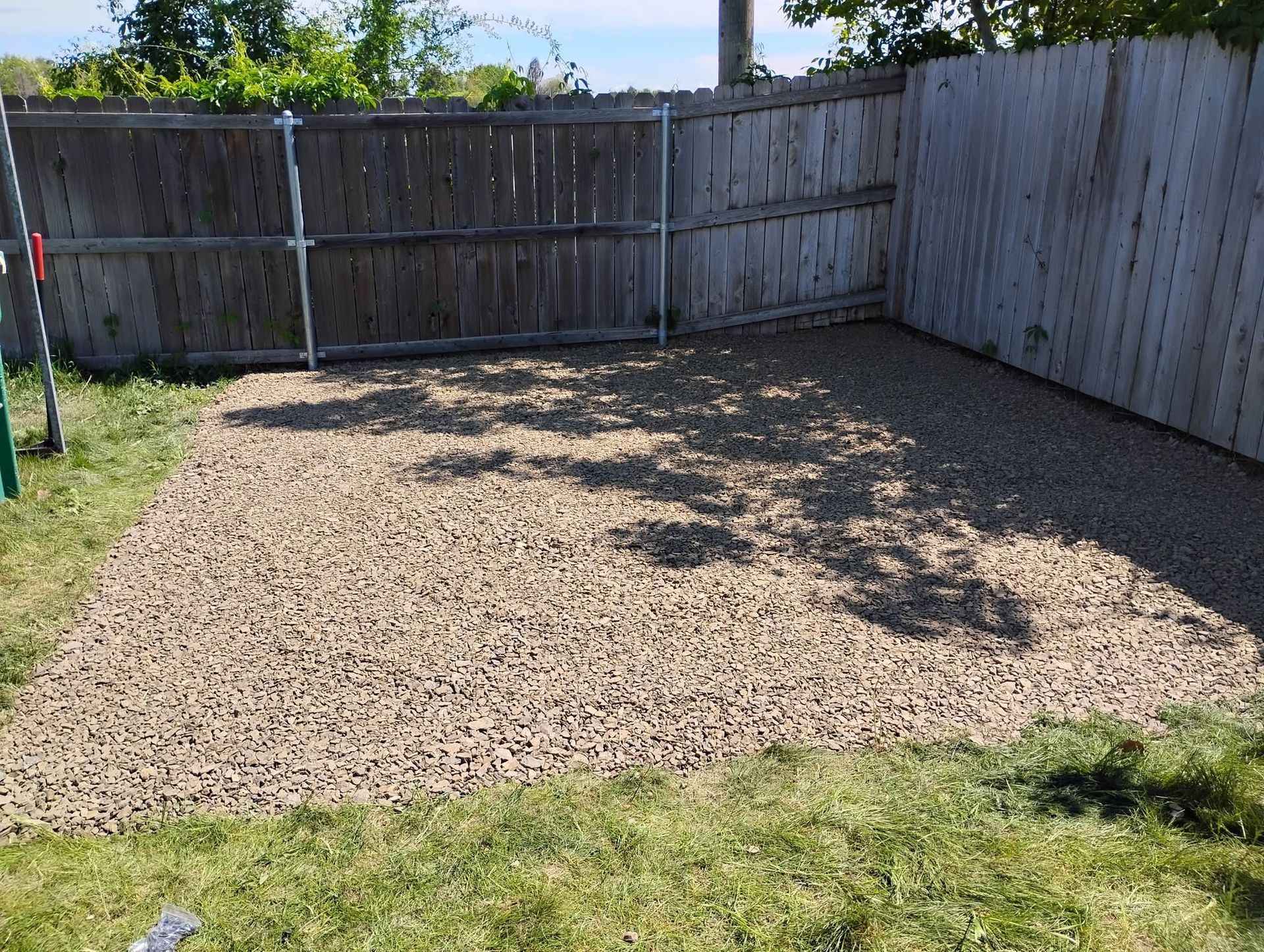 Gravel-covered backyard enclosed by wooden fences, with a small grassy edge and tree shadows.