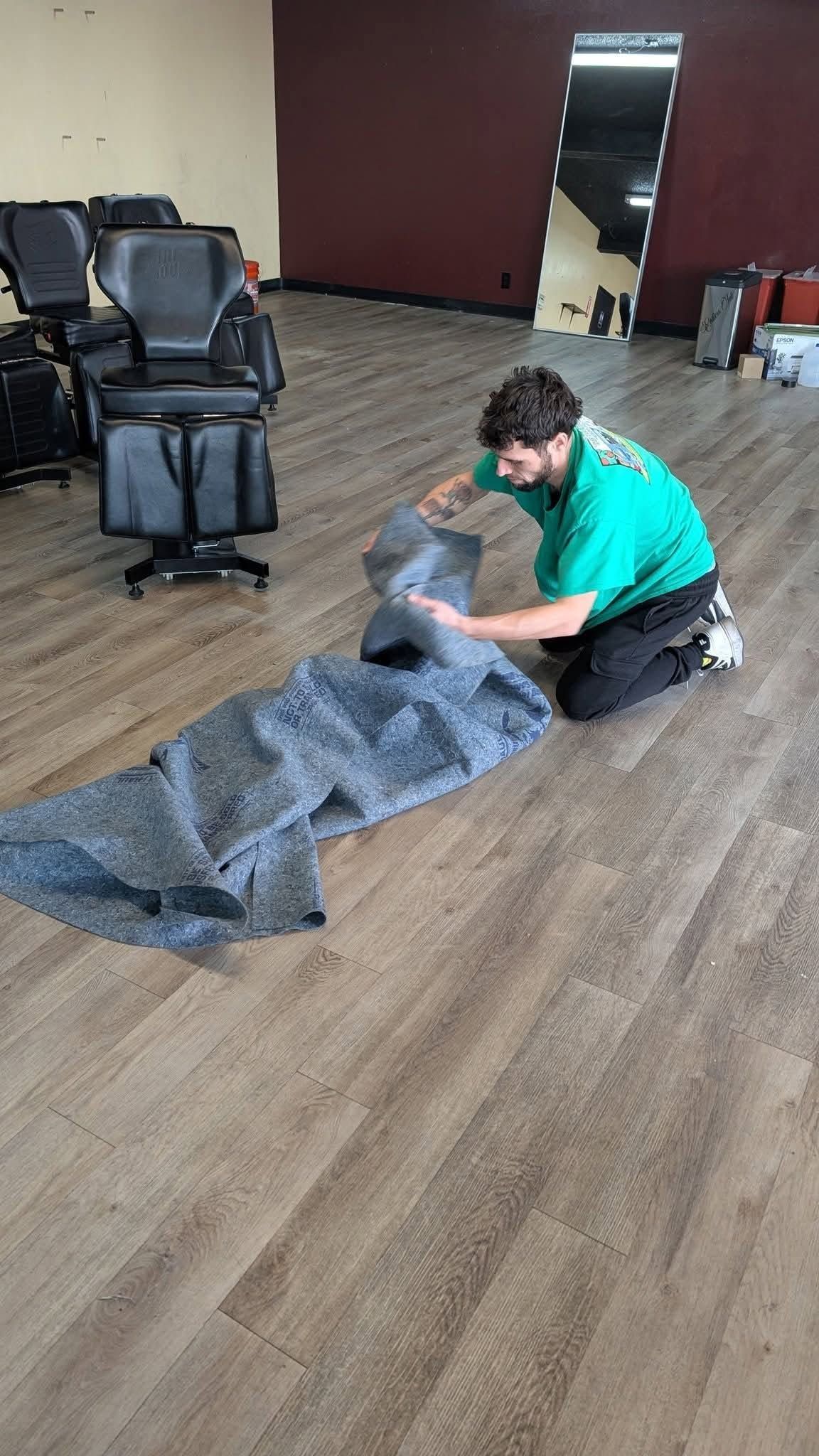 Person kneeling on carpet, folding a gray tarp in a room with stacked chairs and a maroon wall.