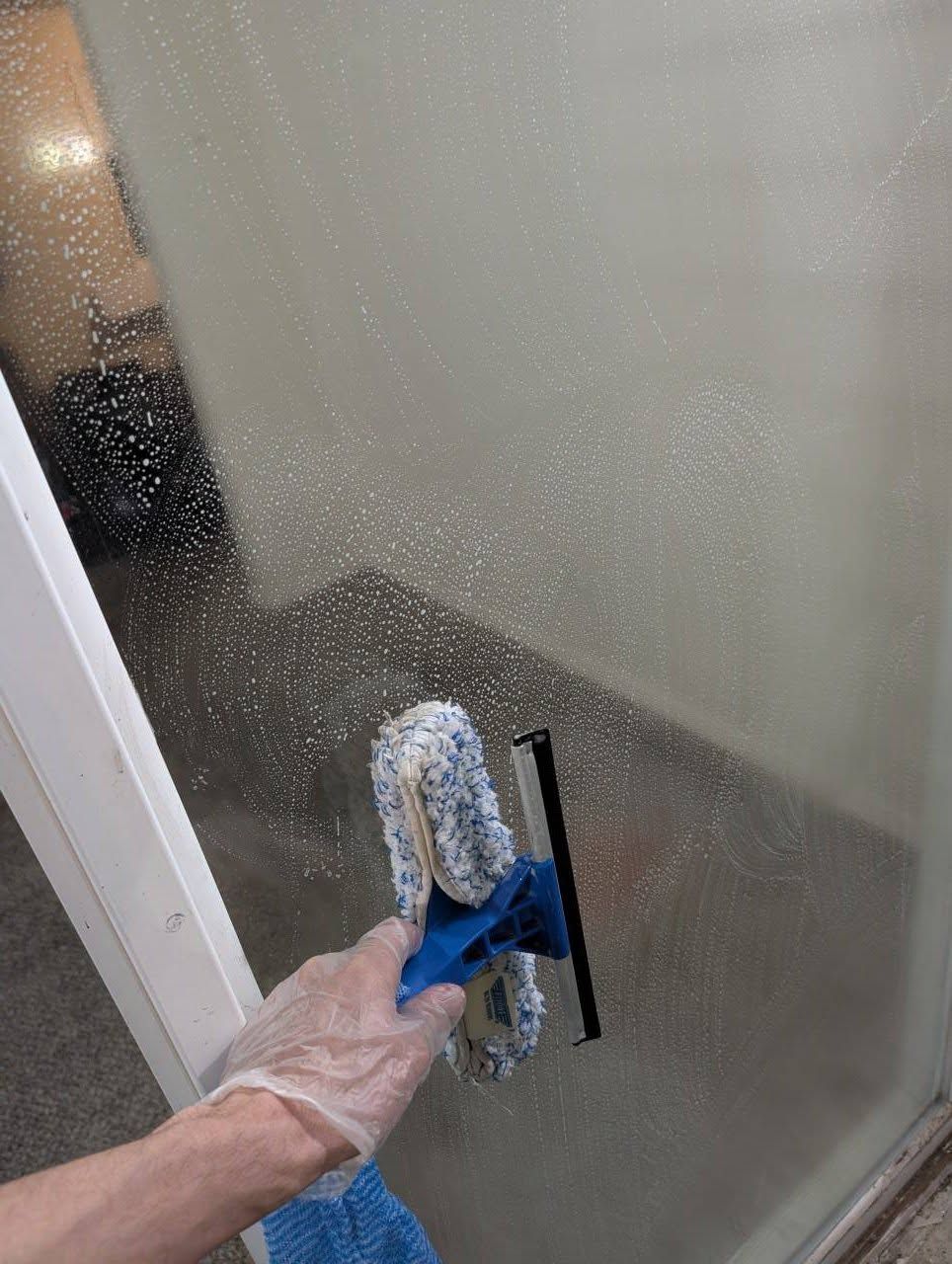 Hand washing a frosted glass shower door with a blue squeegee and cloth