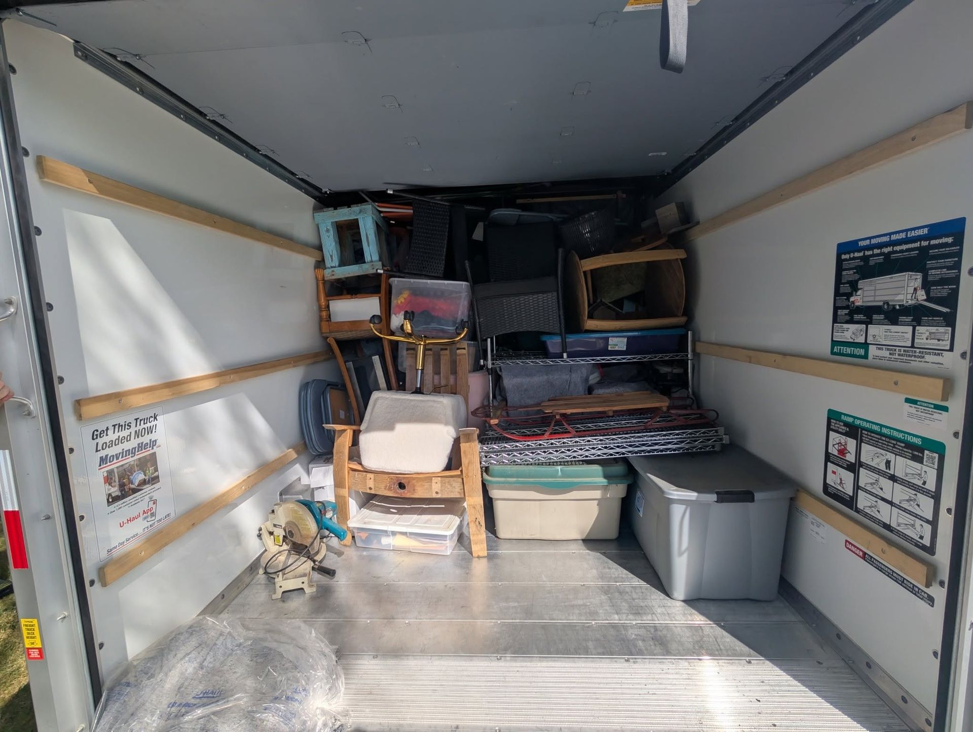 Cluttered cargo trailer interior with stacked boxes, shelves, bins, and a chair near the back