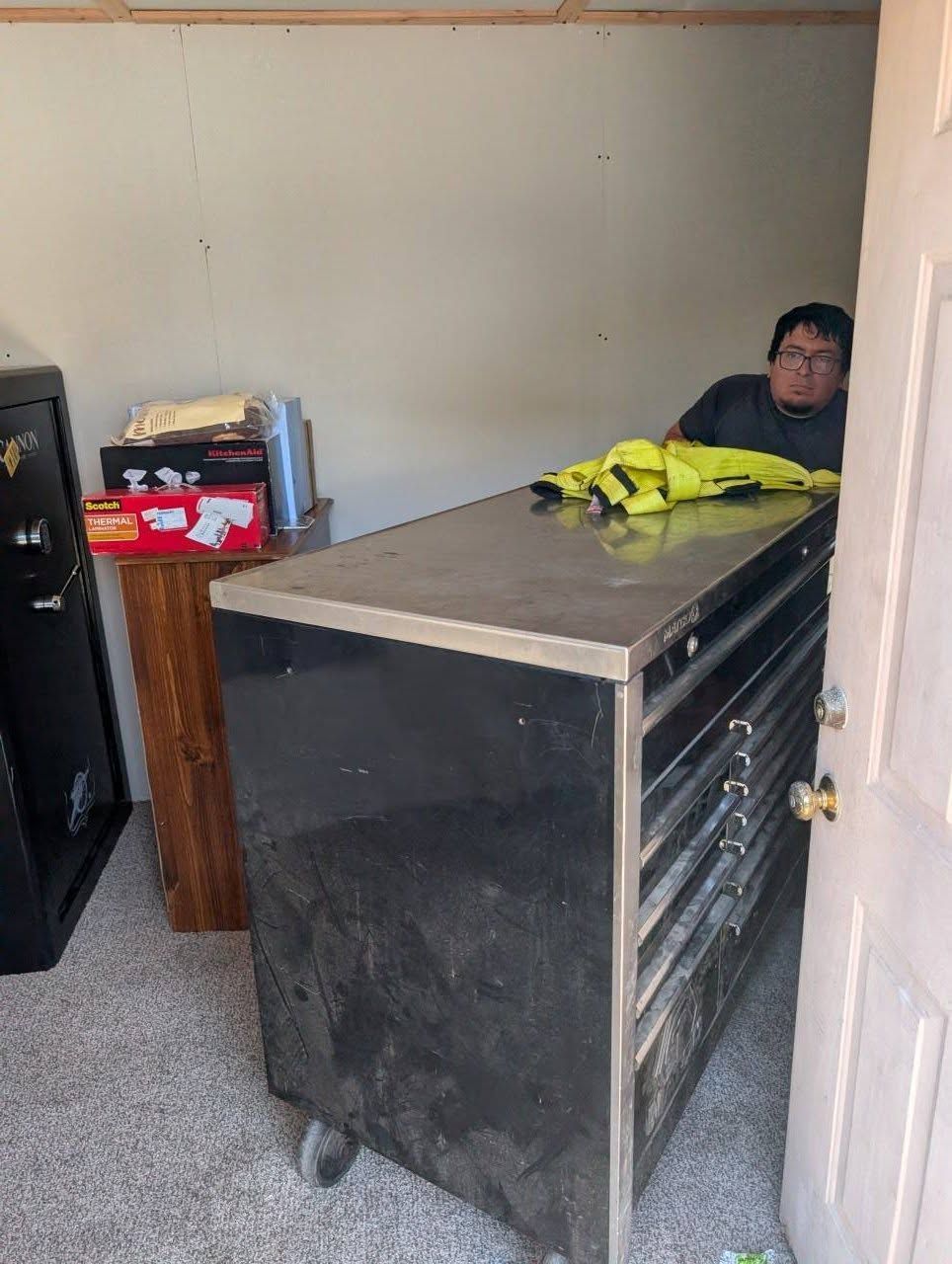 Person sitting behind a large black workbench in a plain room; yellow jacket on top.