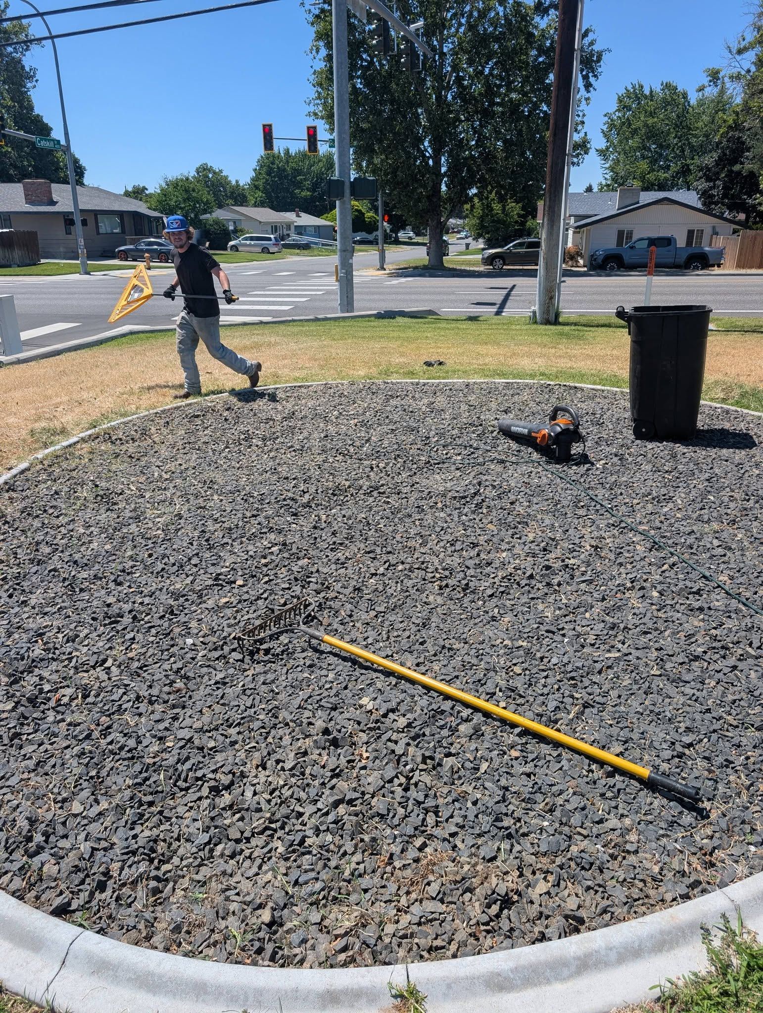 Person walking on gravel beside a trash can and tools in a sunny outdoor work area