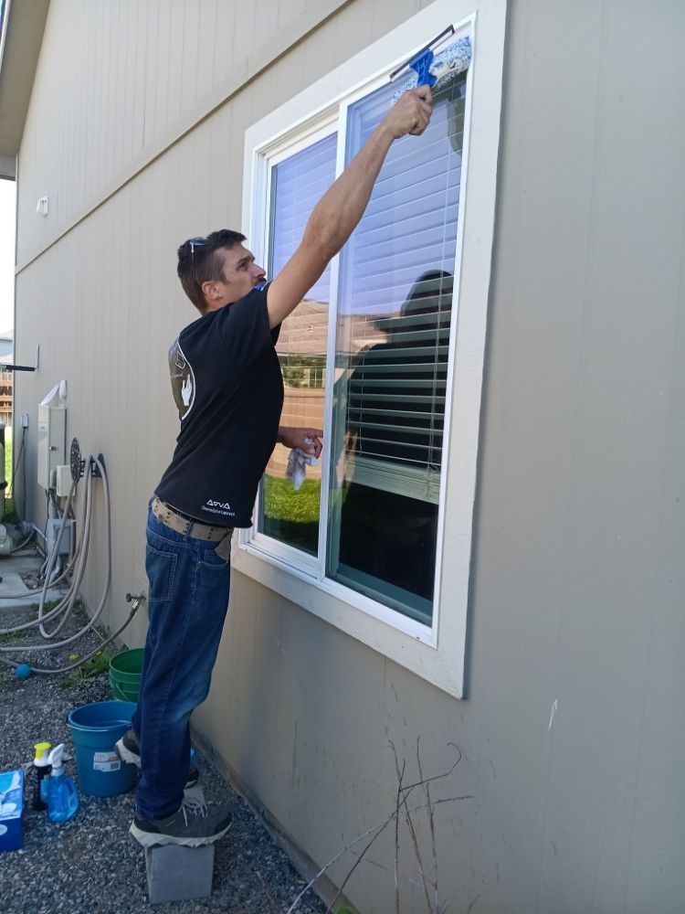 Person cleaning an exterior window with a spray bottle and cloth beside a house wall