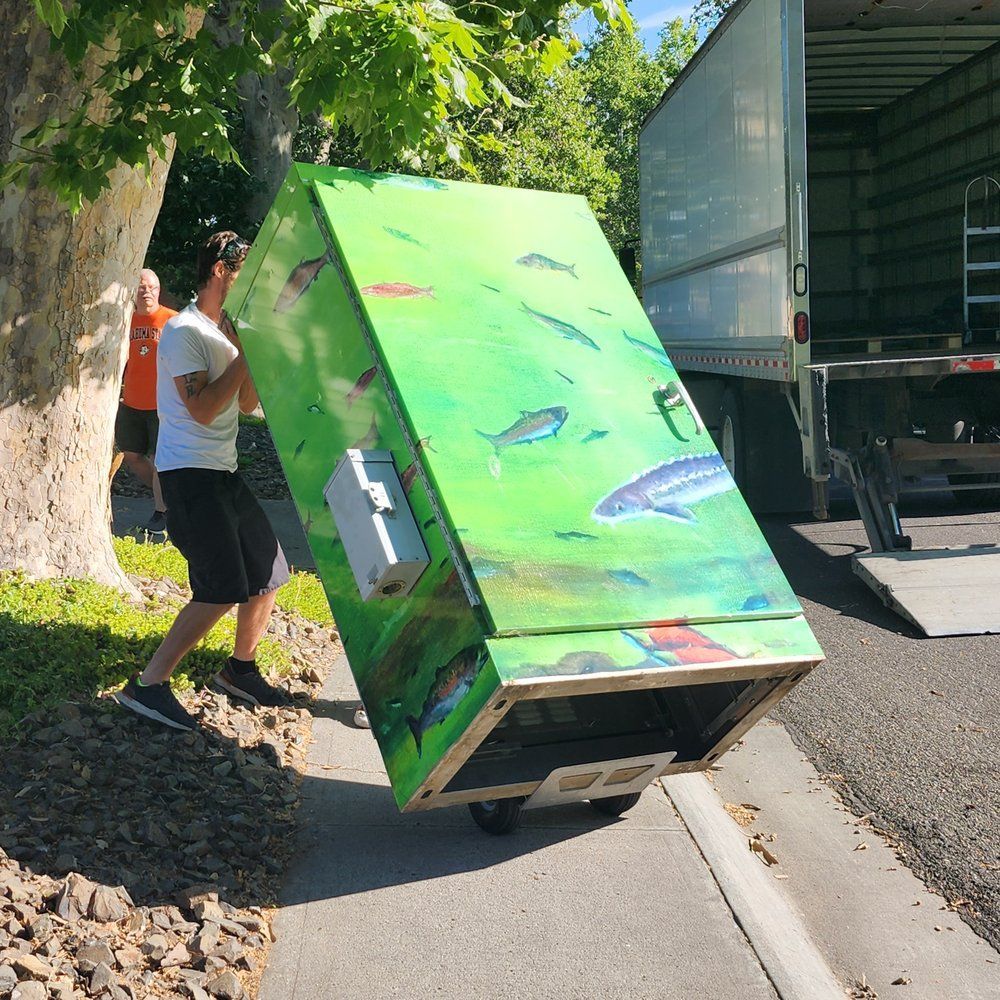 Person pushing a bright green appliance on a hand truck beside a truck and tree-lined roadside