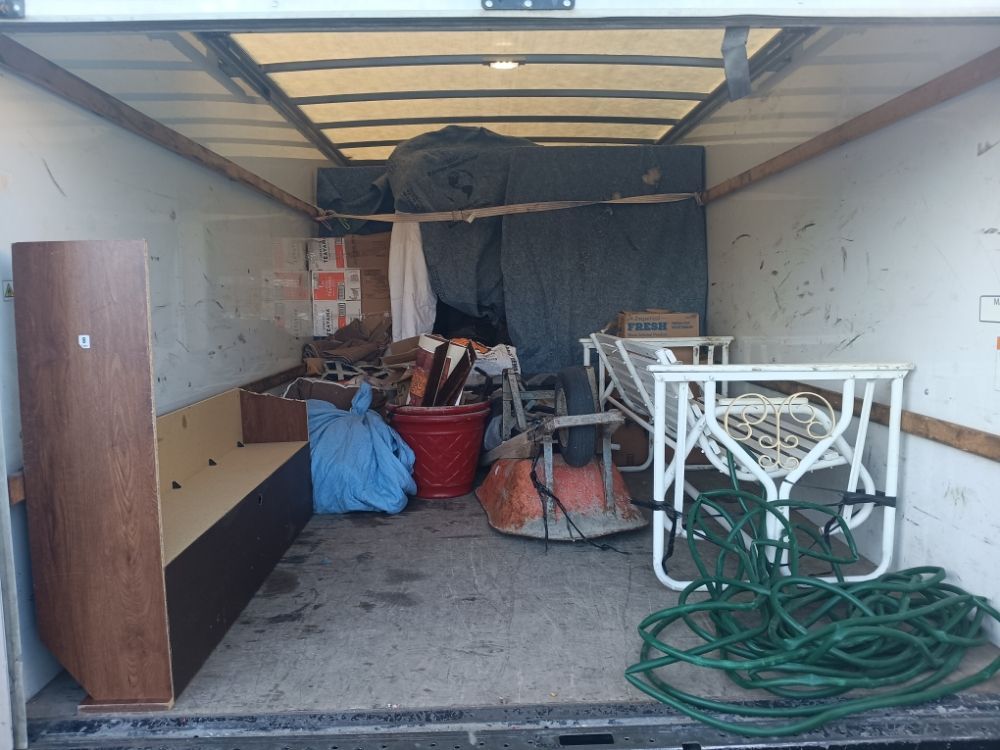Cluttered trailer interior with furniture, boxes, and a coiled green hose near the open rear door