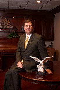 Man standing in an office beside a desk with a winged eagle sculpture, wearing a suit and tie