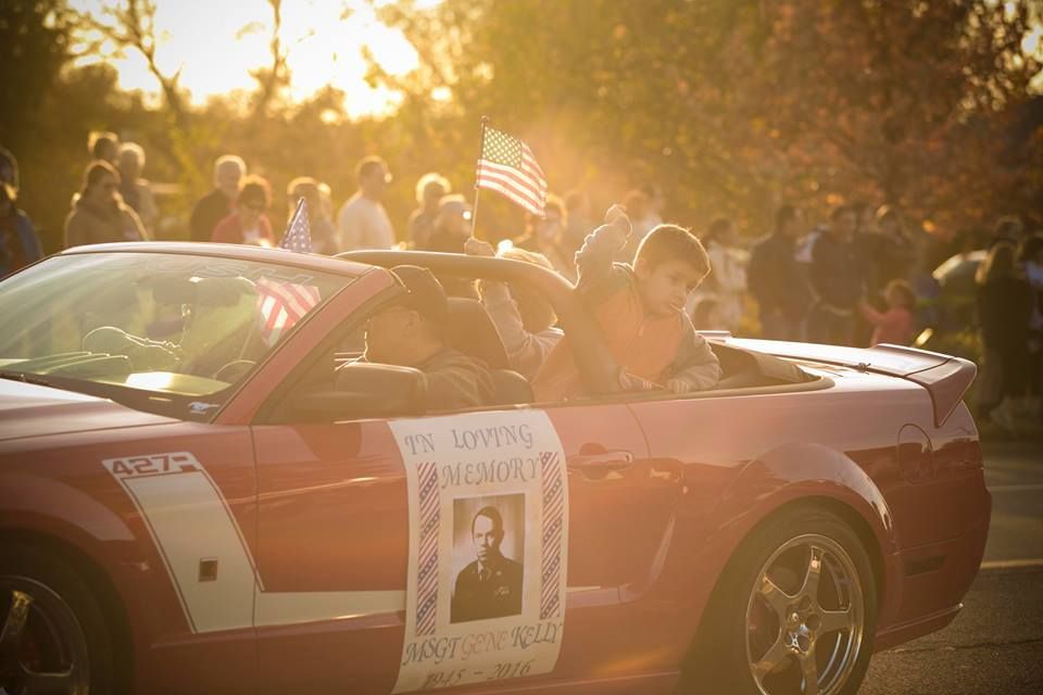 Red convertible in a sunlit parade crowd, with a man leaning out and a campaign poster on the door