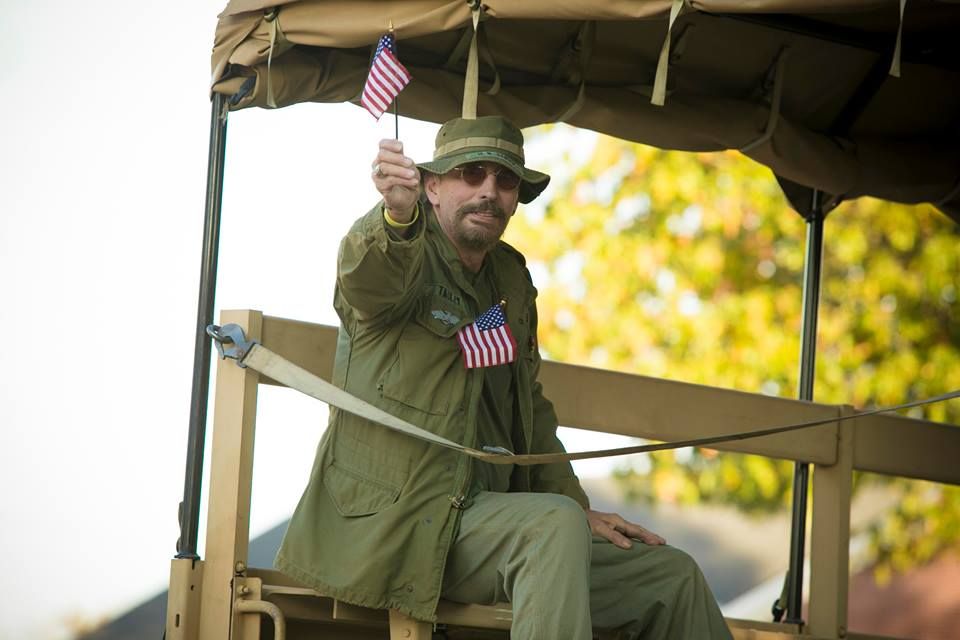 Man in green rain gear on a military-style vehicle, holding up small American flags under a canvas canopy
