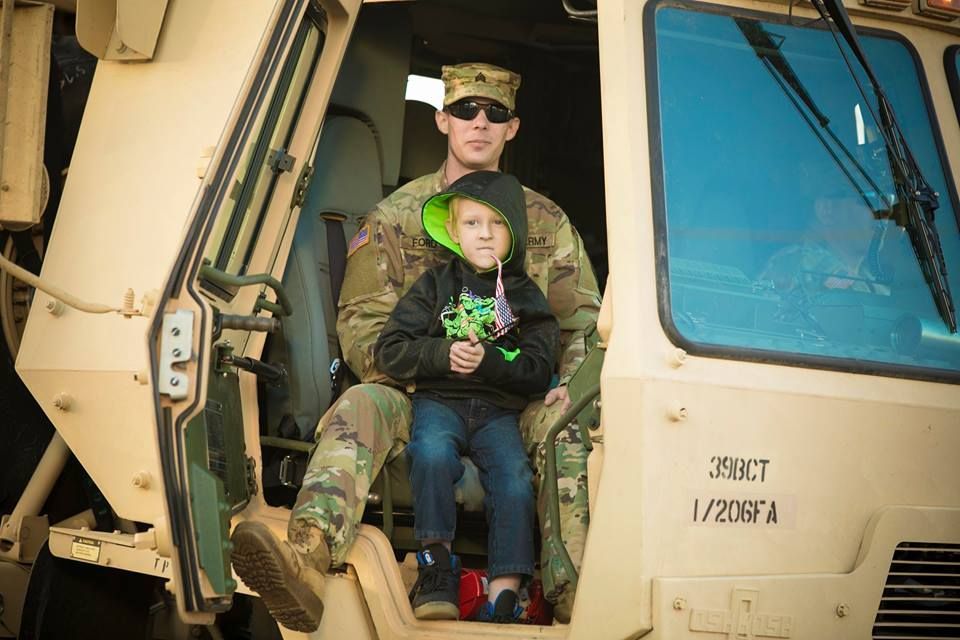 Soldier holding a child in camouflage on a military vehicle step, both facing the camera