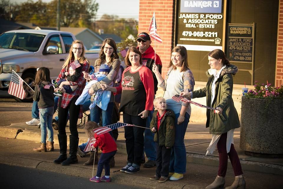 People holding a blue rope at an outdoor community event beside a brick building and parked cars.