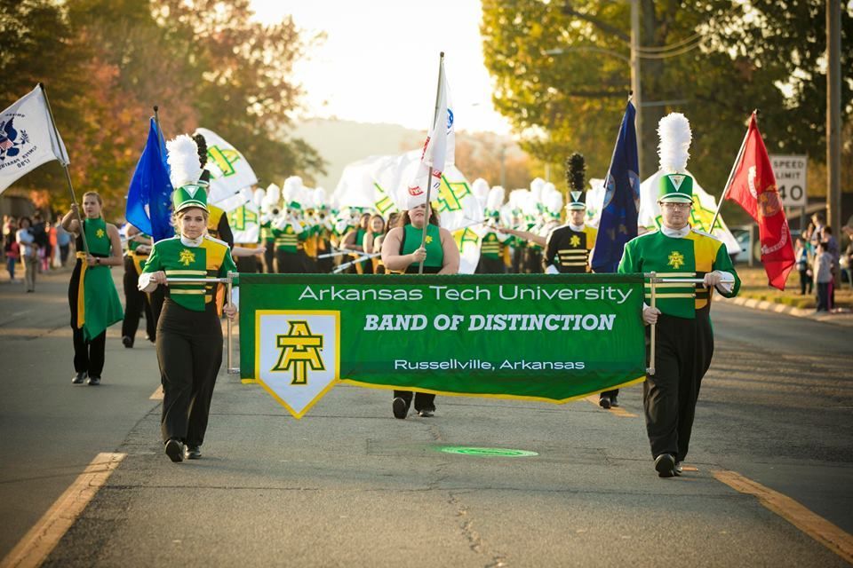 Arkansas Tech University band marching in parade, carrying green banner and flags at sunset.