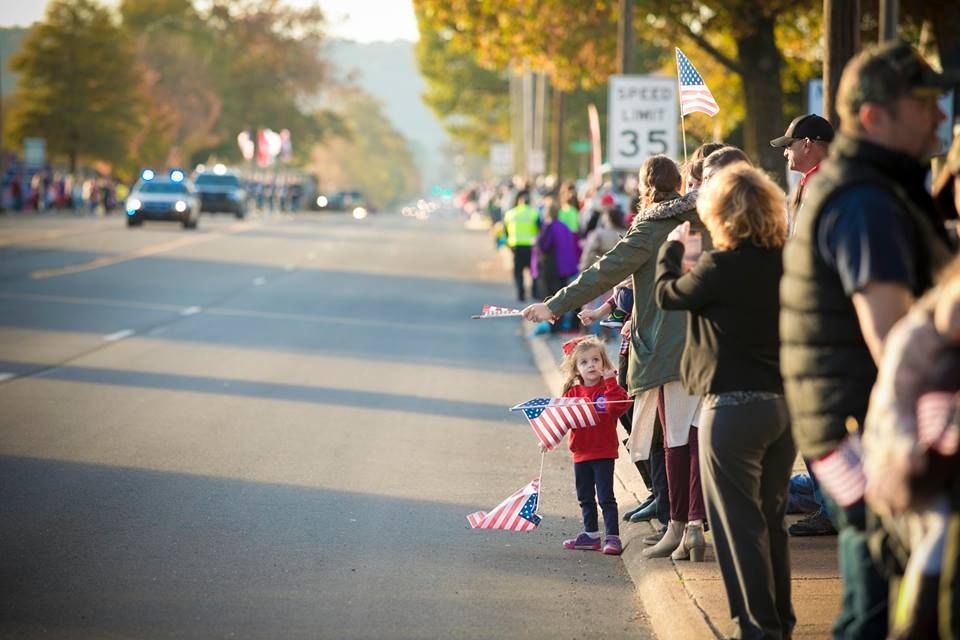 People lining a tree-lined street, waving small American flags during a parade or procession.