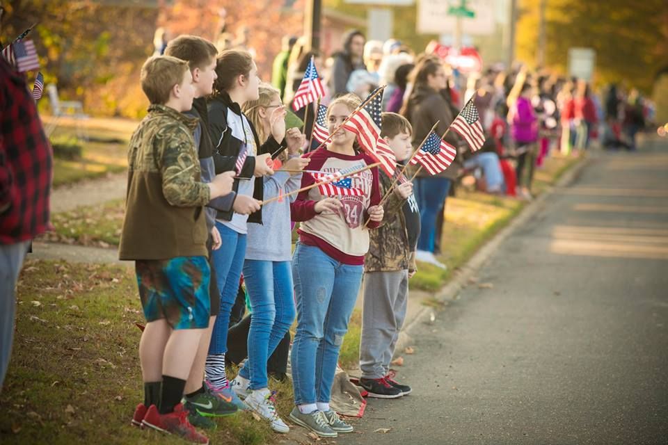 Children holding small American flags line a roadside during a sunny outdoor event.